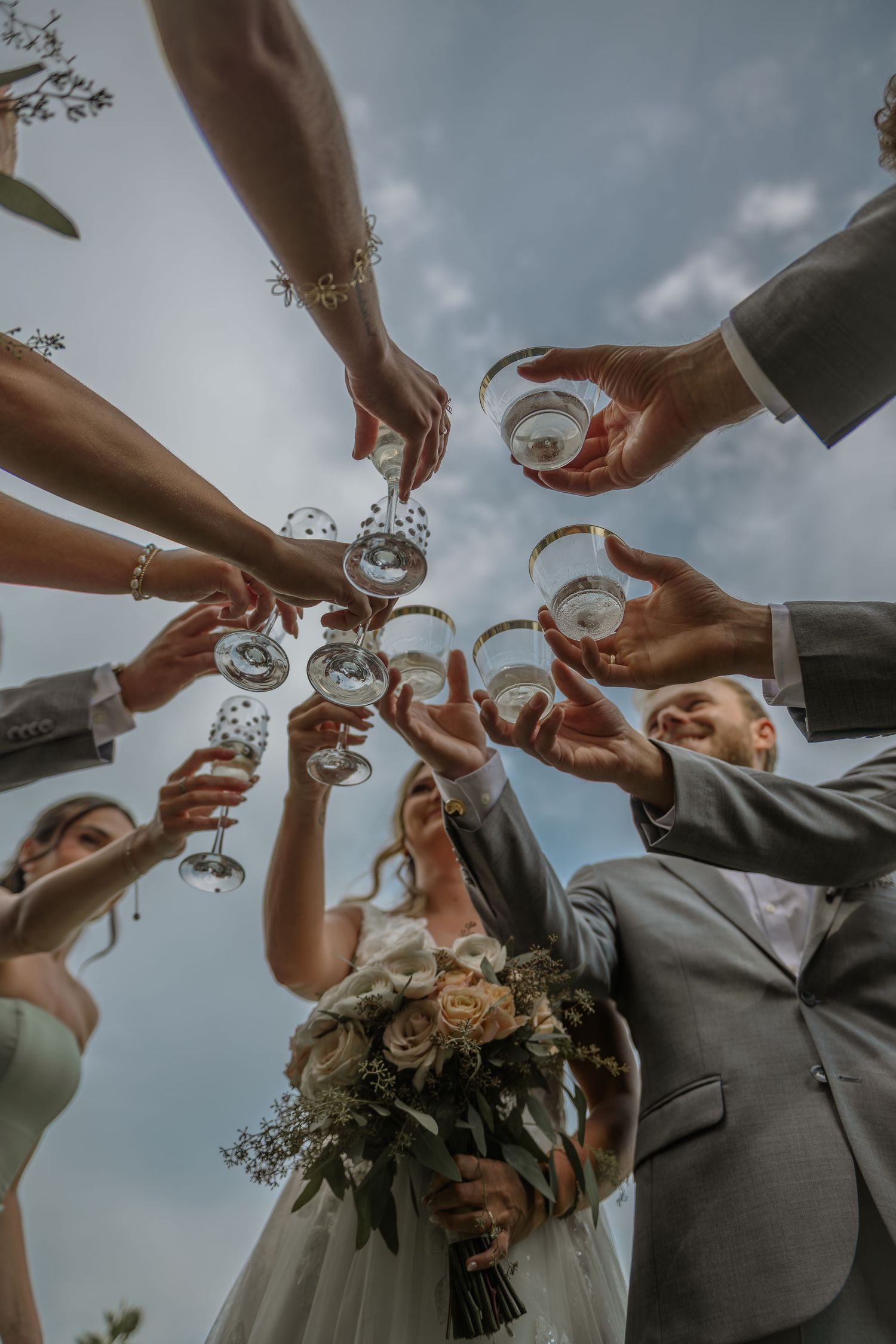 Wedding party toasting with champagne glasses against a cloudy sky.