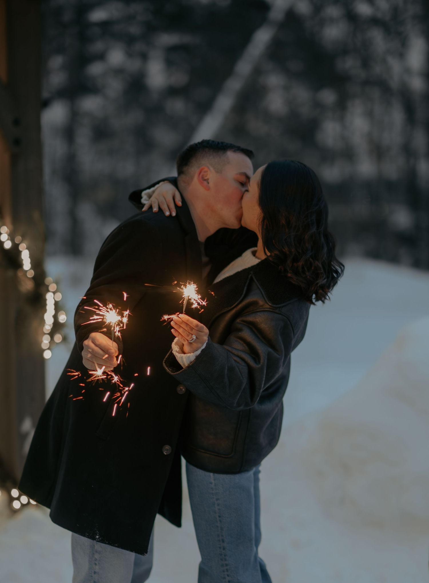 Couple holding sparklers in snowy winter setting with twinkling lights.