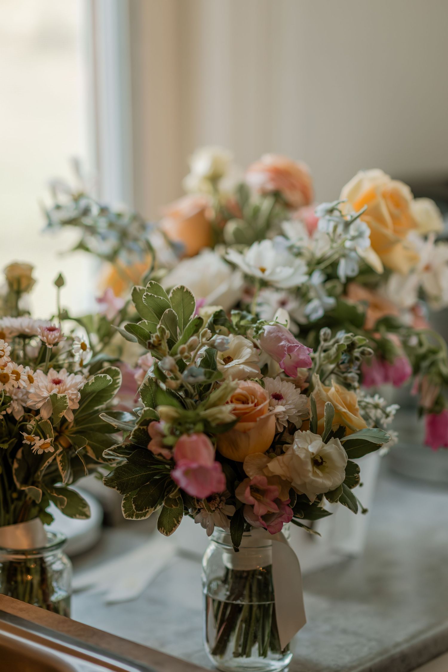 A row of elegant floral centerpieces with peach roses and mixed flowers in glass vases displayed on a windowsill.
