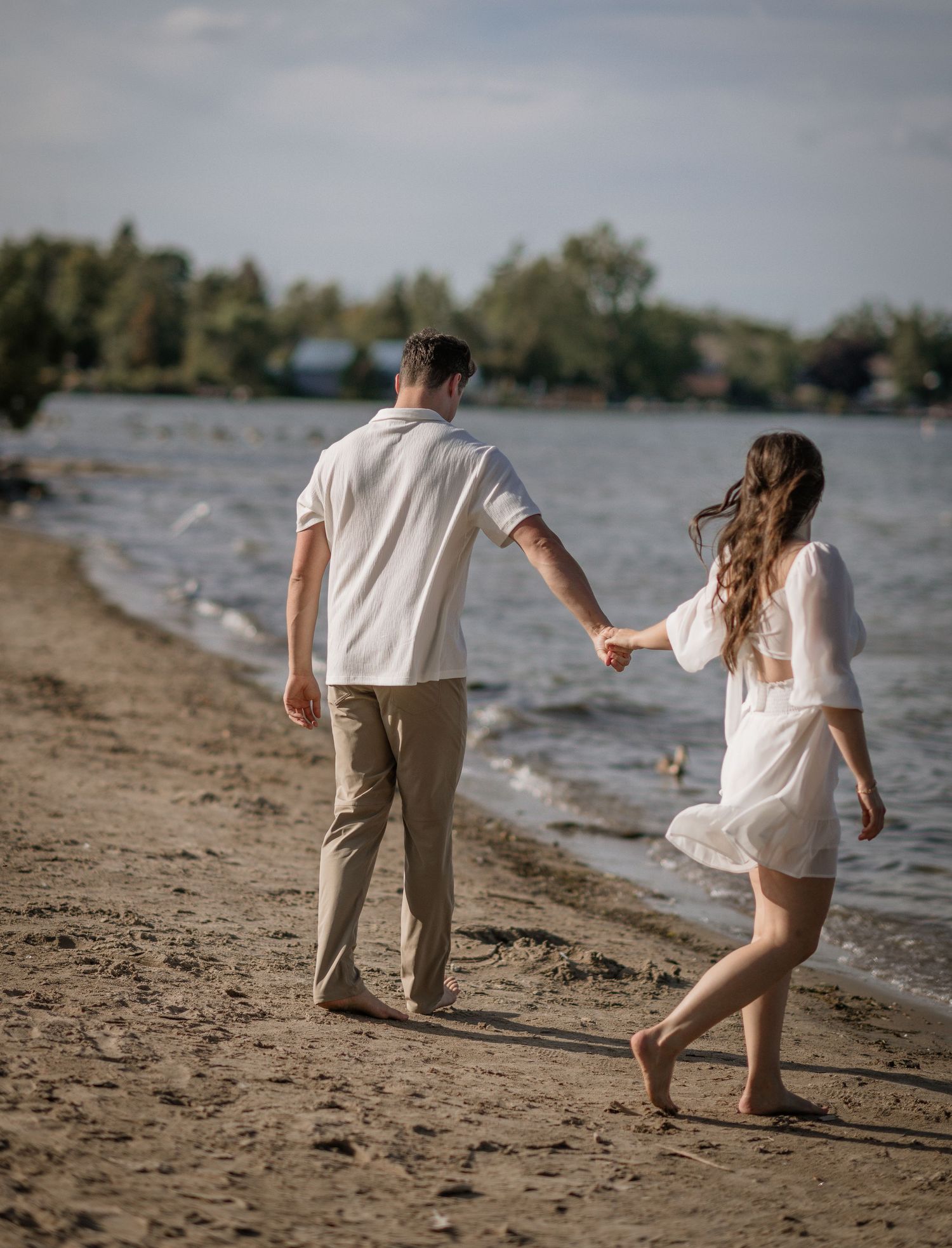 A couple holds hands while walking barefoot along a lakeside beach.