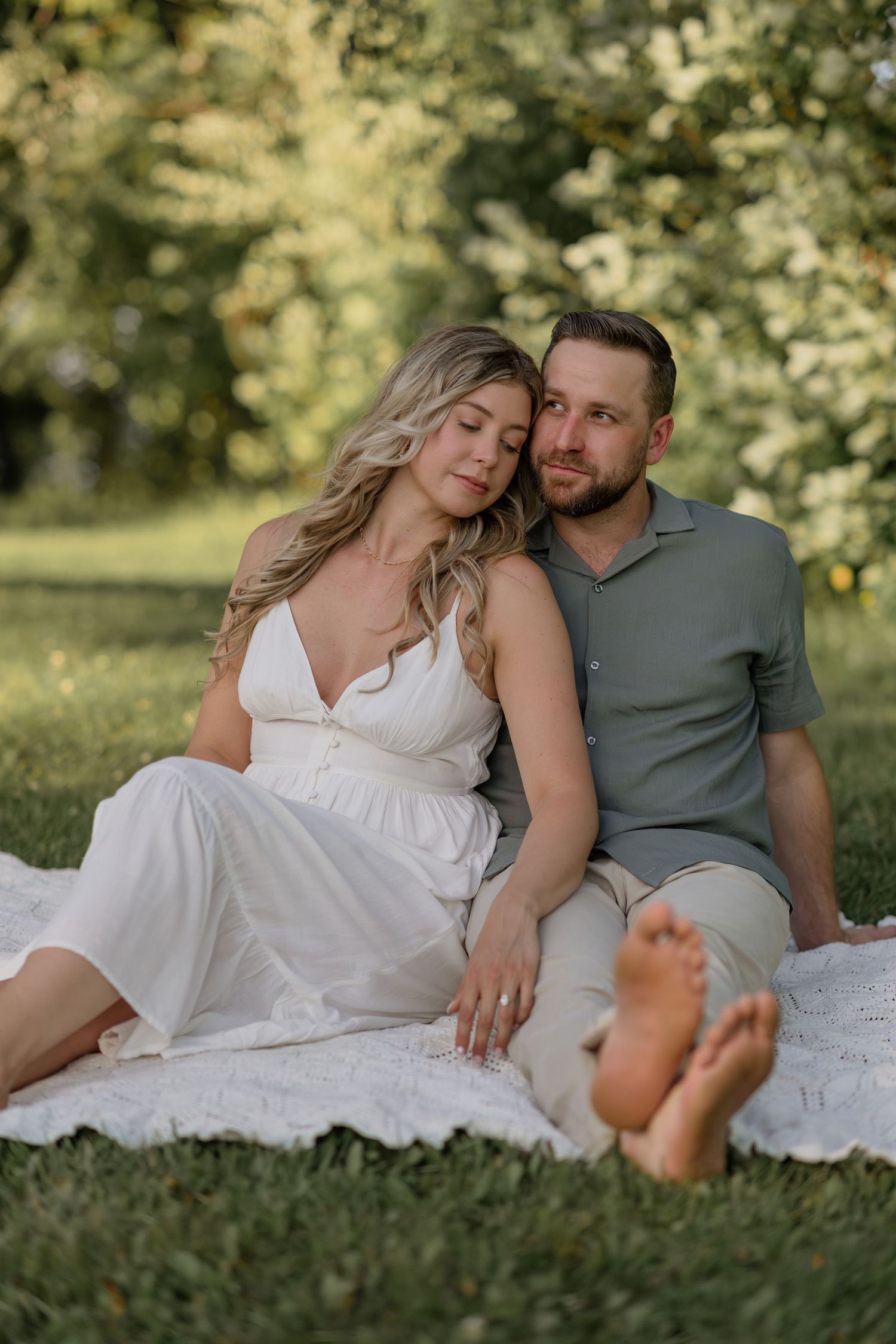Couple relaxes on white blanket in sunny garden setting.