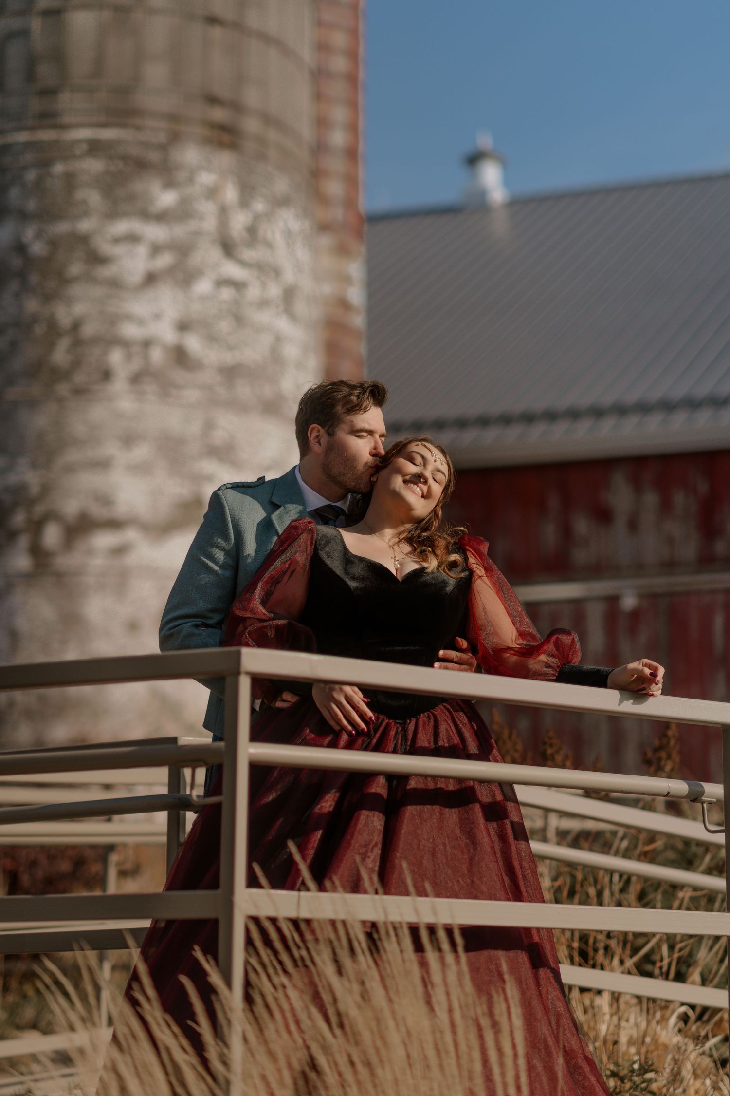 Romantic couple in vintage attire embrace on rustic farm balcony with red barn and silo in background.