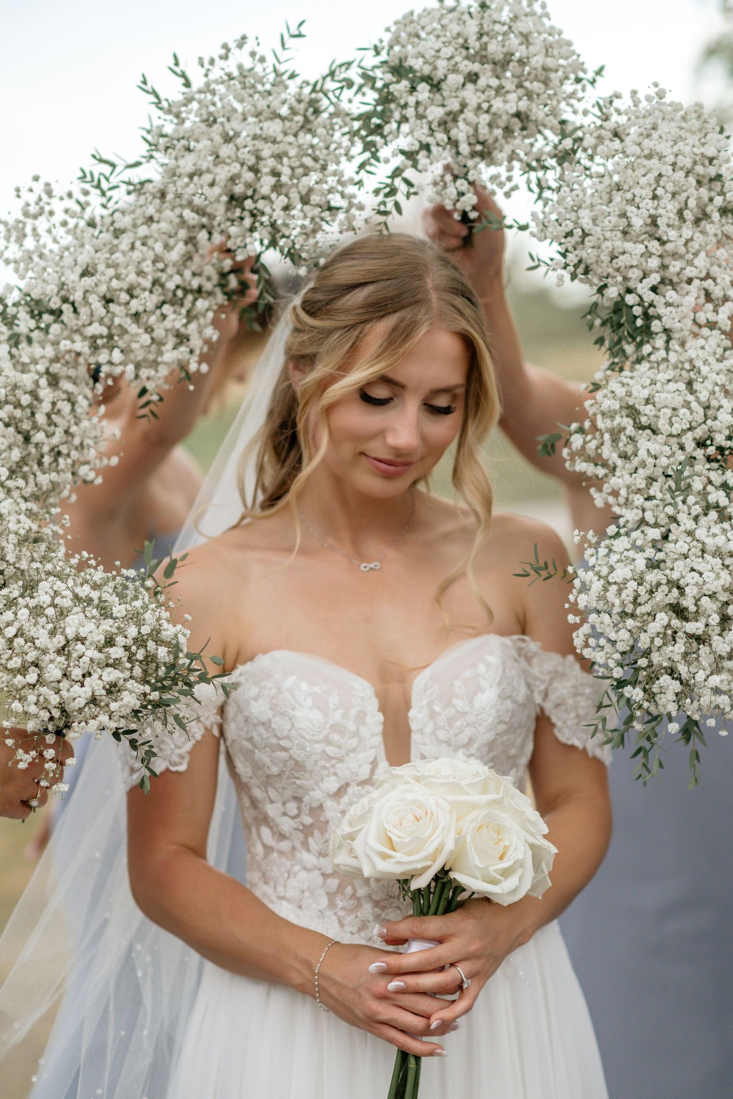 A bride holds white roses while surrounded by baby's breath flowers and wearing a lace wedding gown.