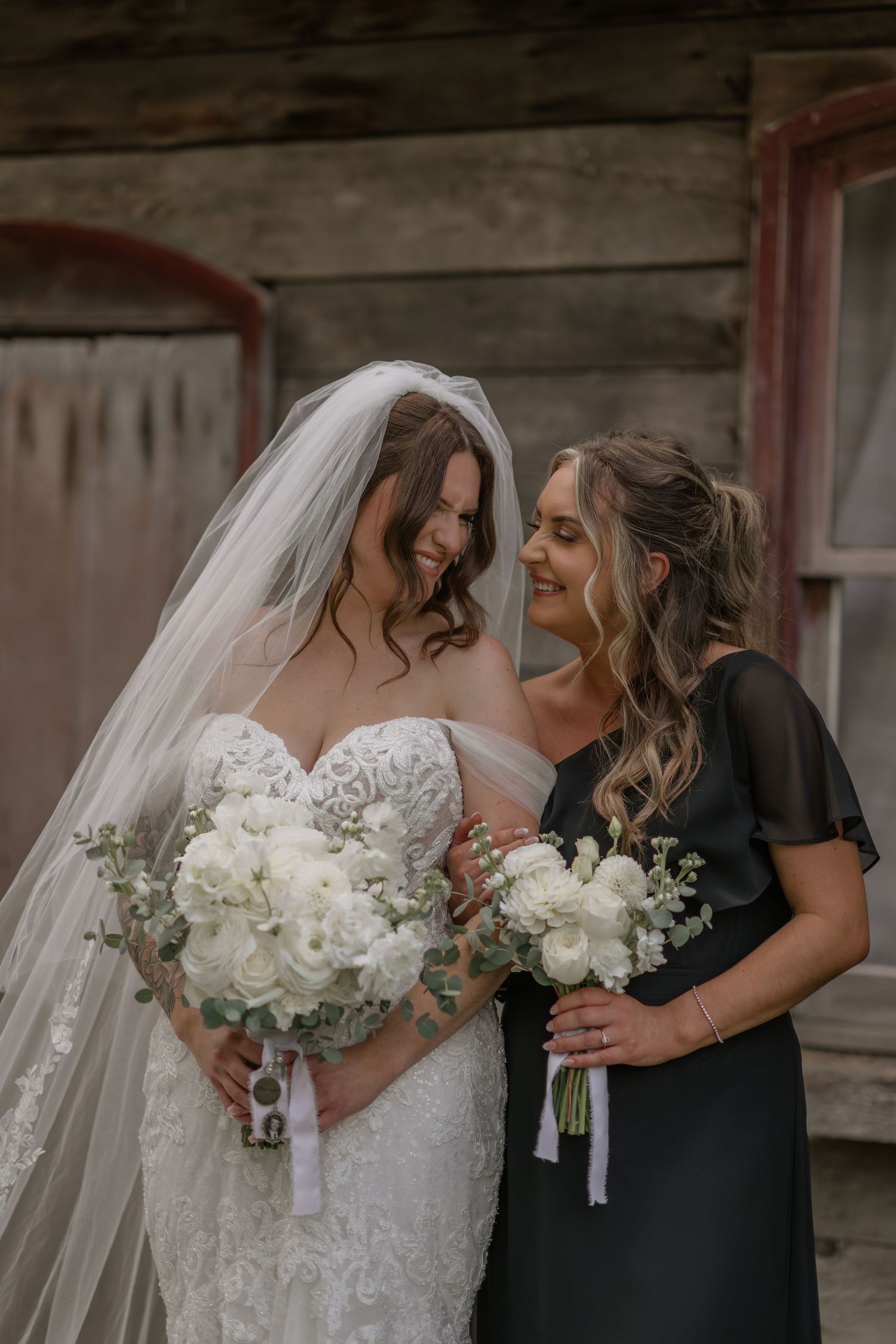 Bridesmaids in black dresses holding white bouquets pose with bride in front of rustic barn.