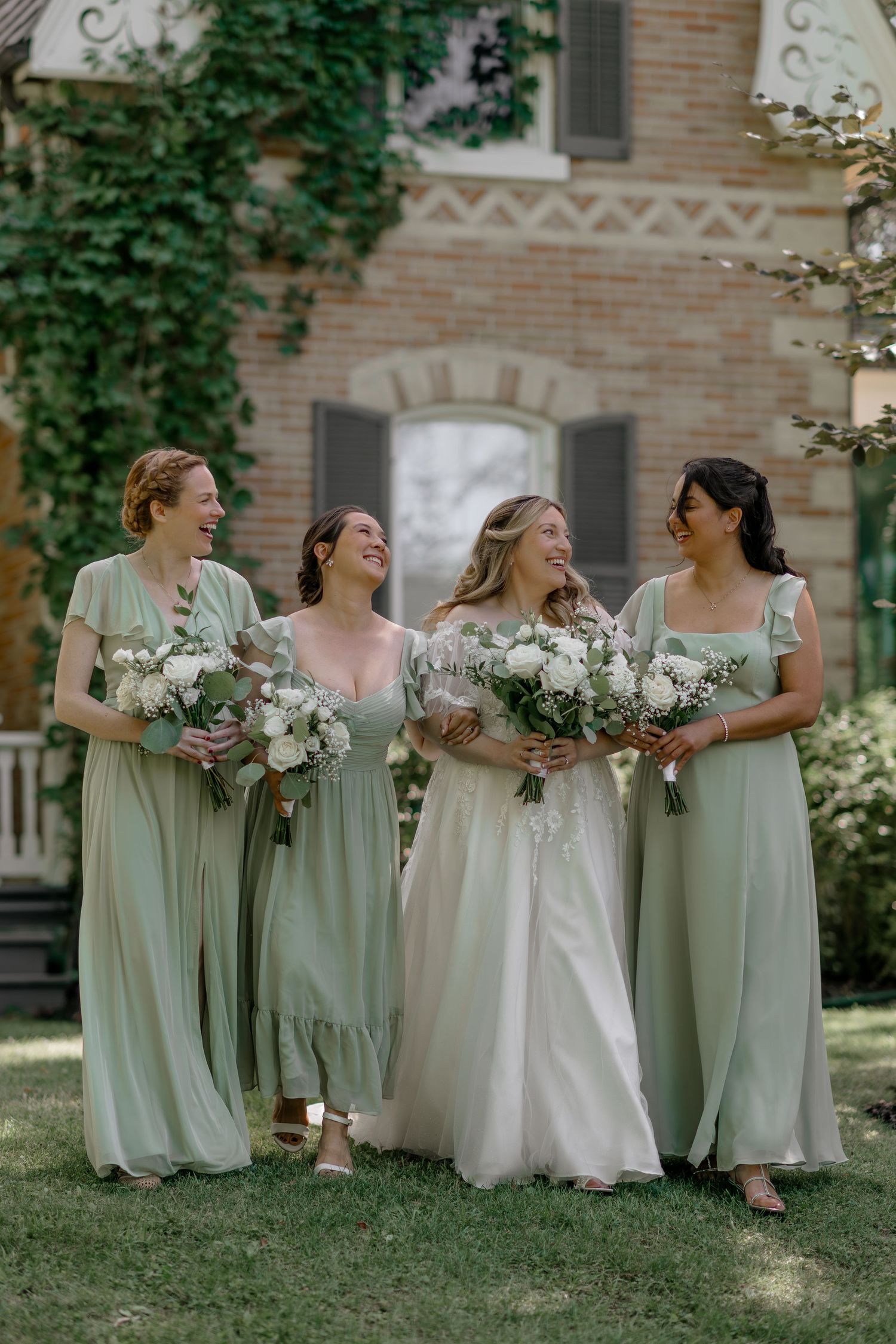 Bridesmaids in sage green dresses laugh together on grass near ivy-covered building.