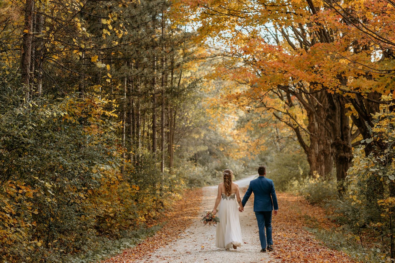 A couple walks down a tree-lined path surrounded by vibrant autumn foliage in golden afternoon light.