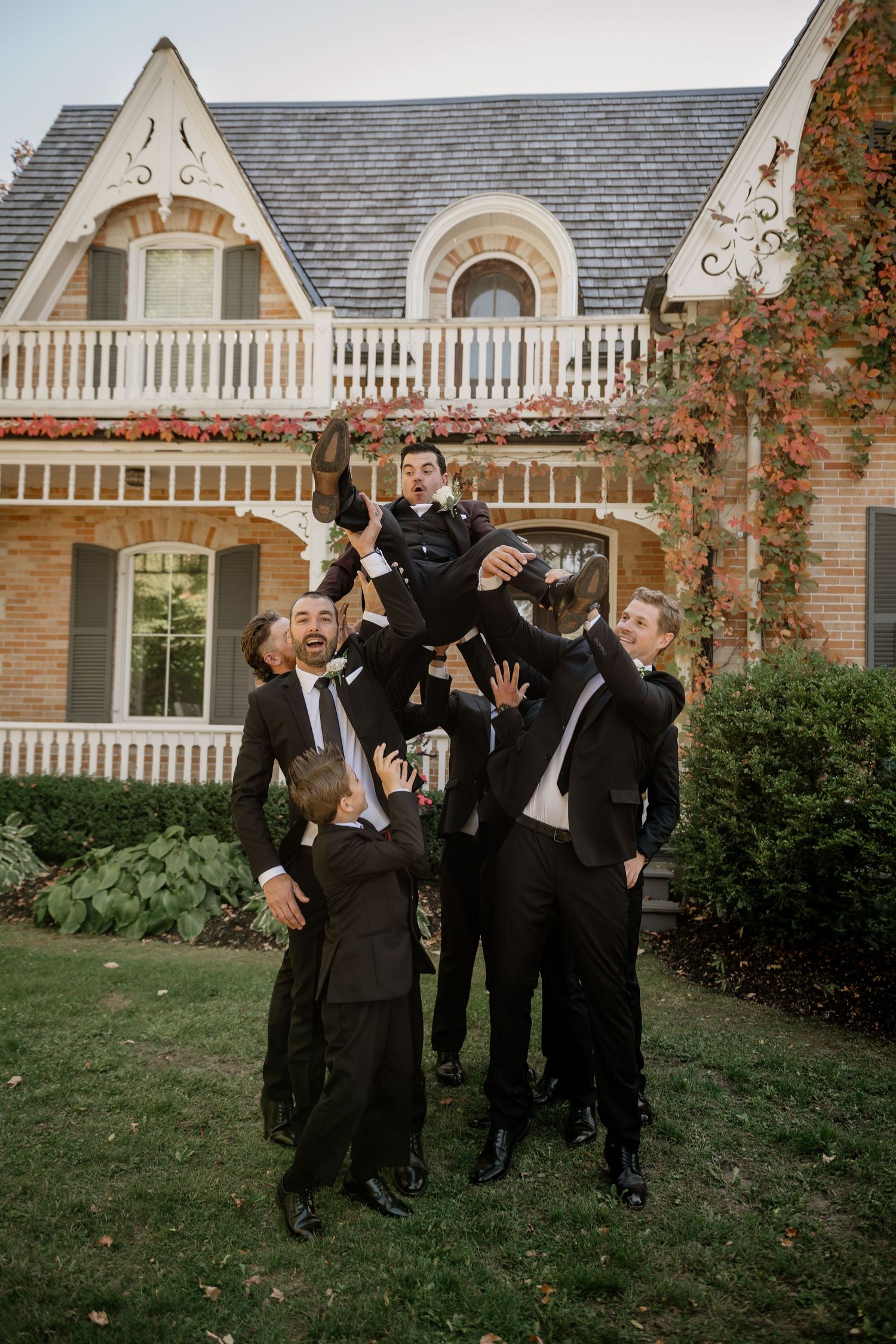 A group in formal black attire poses playfully on a lawn in front of an elegant stone house with arched details.