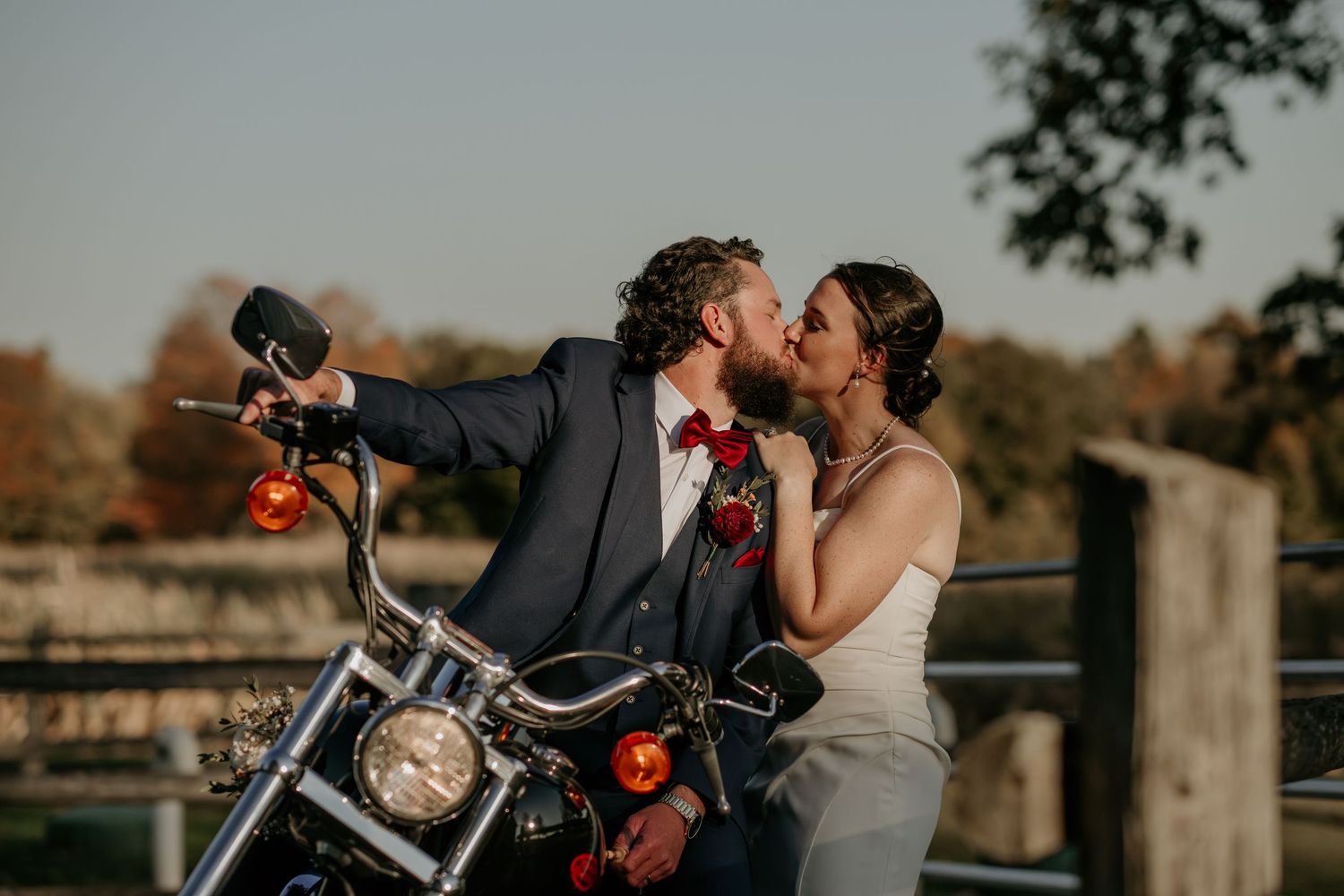 A romantic couple share a tender moment while posing with a motorcycle during their wedding photoshoot at sunset.