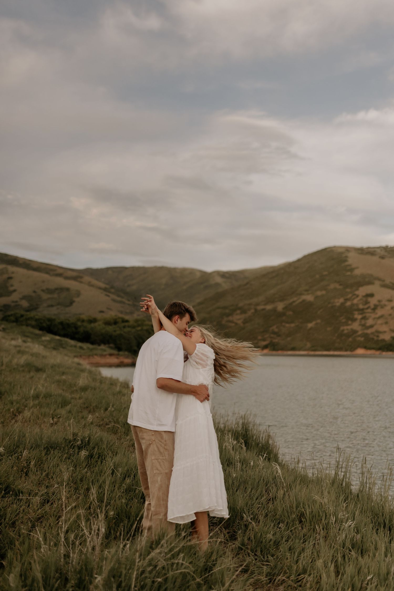 Couple dancing together on a grassy hillside overlooking a serene lake during golden hour.