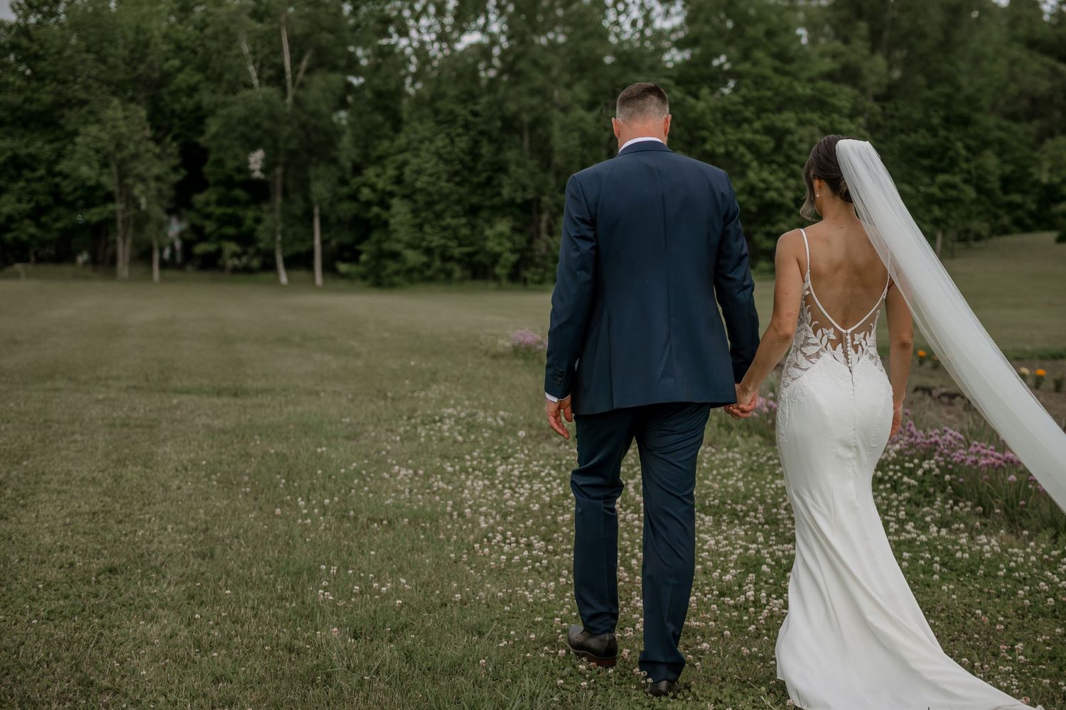 Newlywed couple walking together on grassy field with bride in fitted white dress and groom in navy suit.