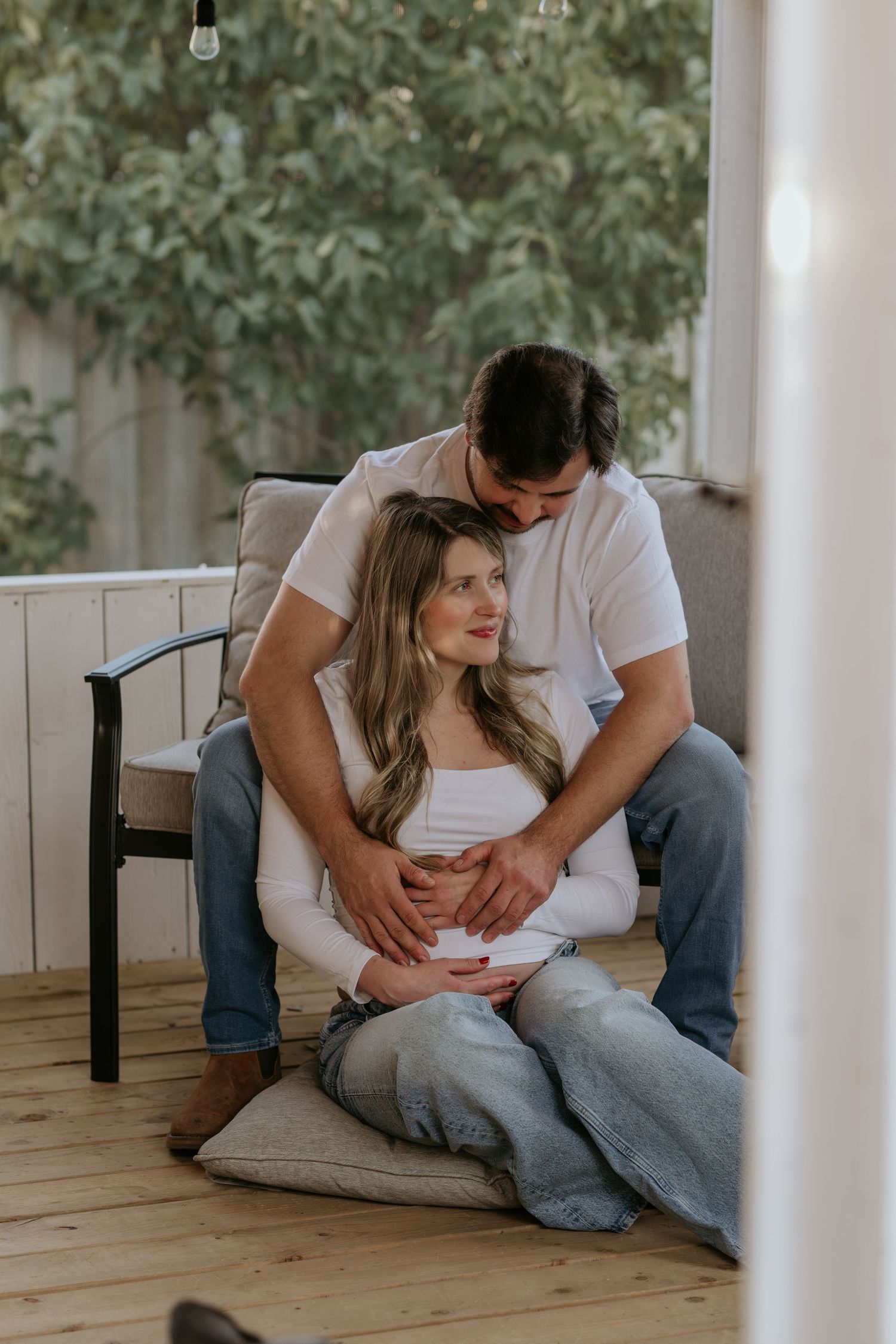 Romantic maternity photos on wooden deck with white pillars and greenery in background.