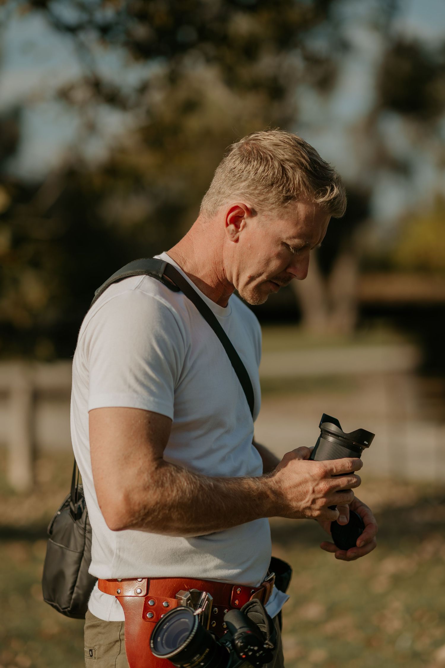 Person in white t-shirt checking mobile phone while holding camera outdoors during golden hour.