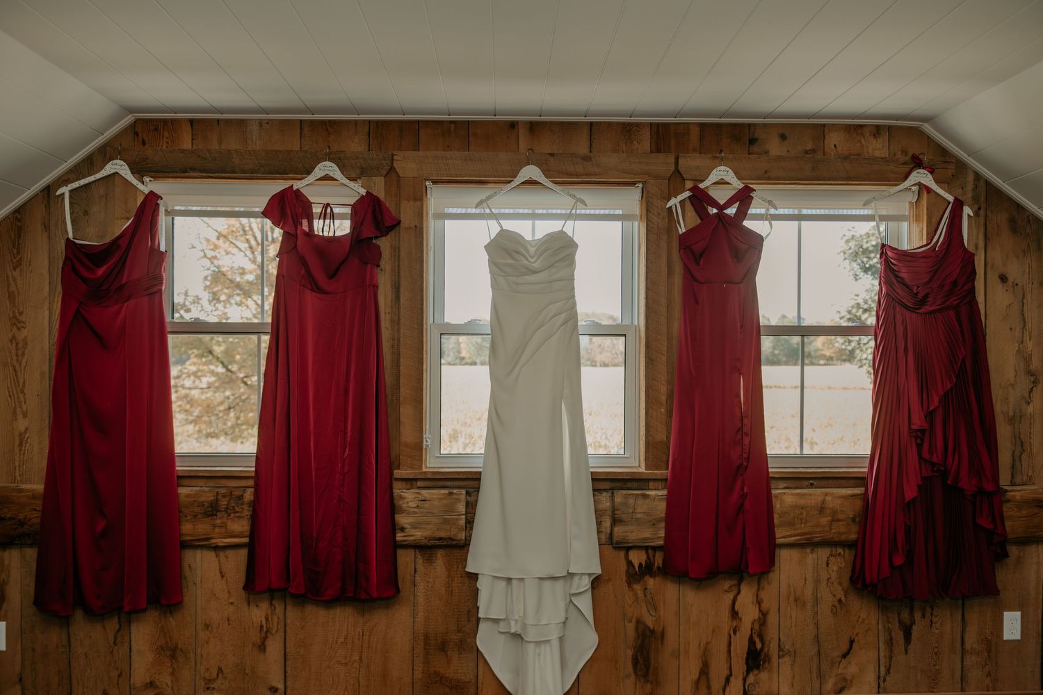 Wedding party dresses in burgundy red and white hang on wooden wall hooks in rustic venue with natural lighting.