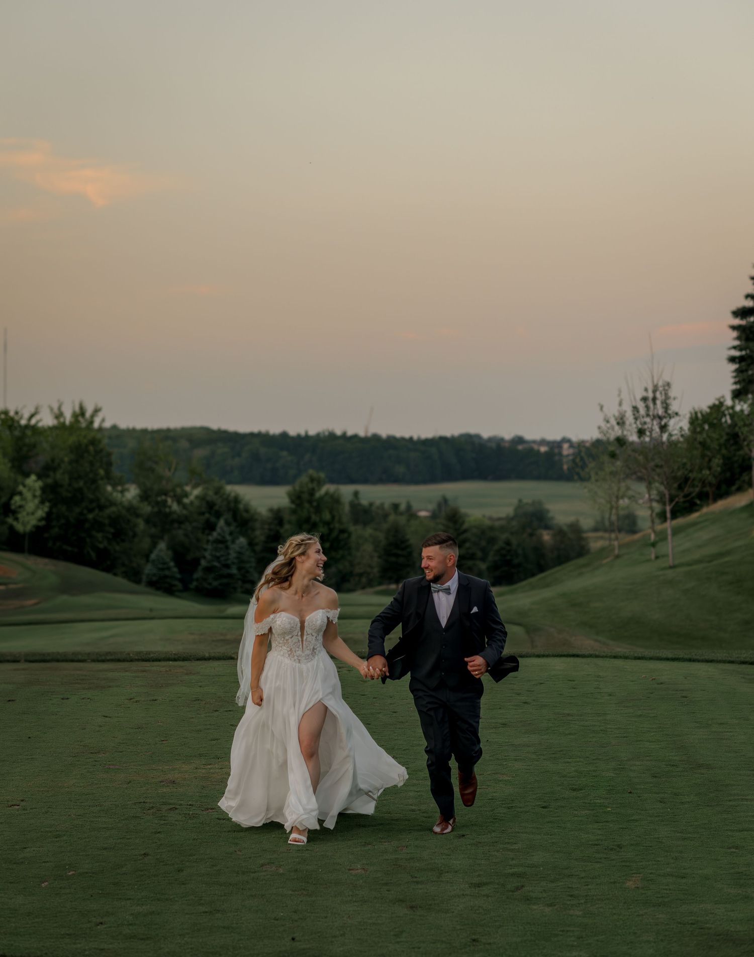 Wedding couple runs playfully across green hillside at sunset.