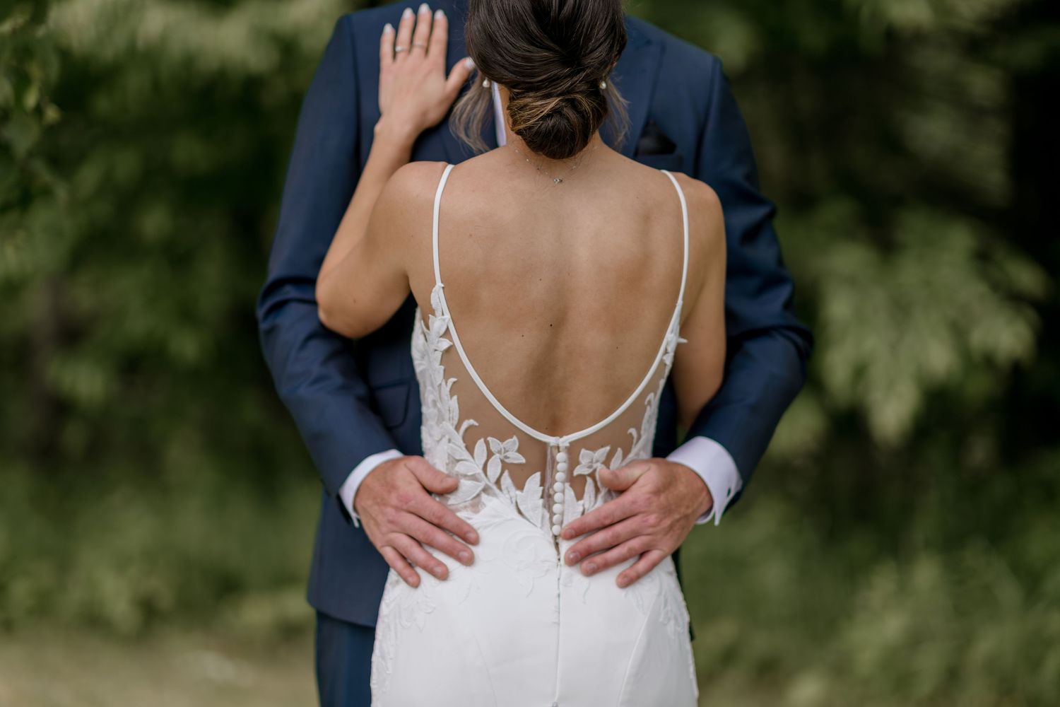 Close-up of a bride in a backless white lace wedding dress with spaghetti straps being embraced from behind.