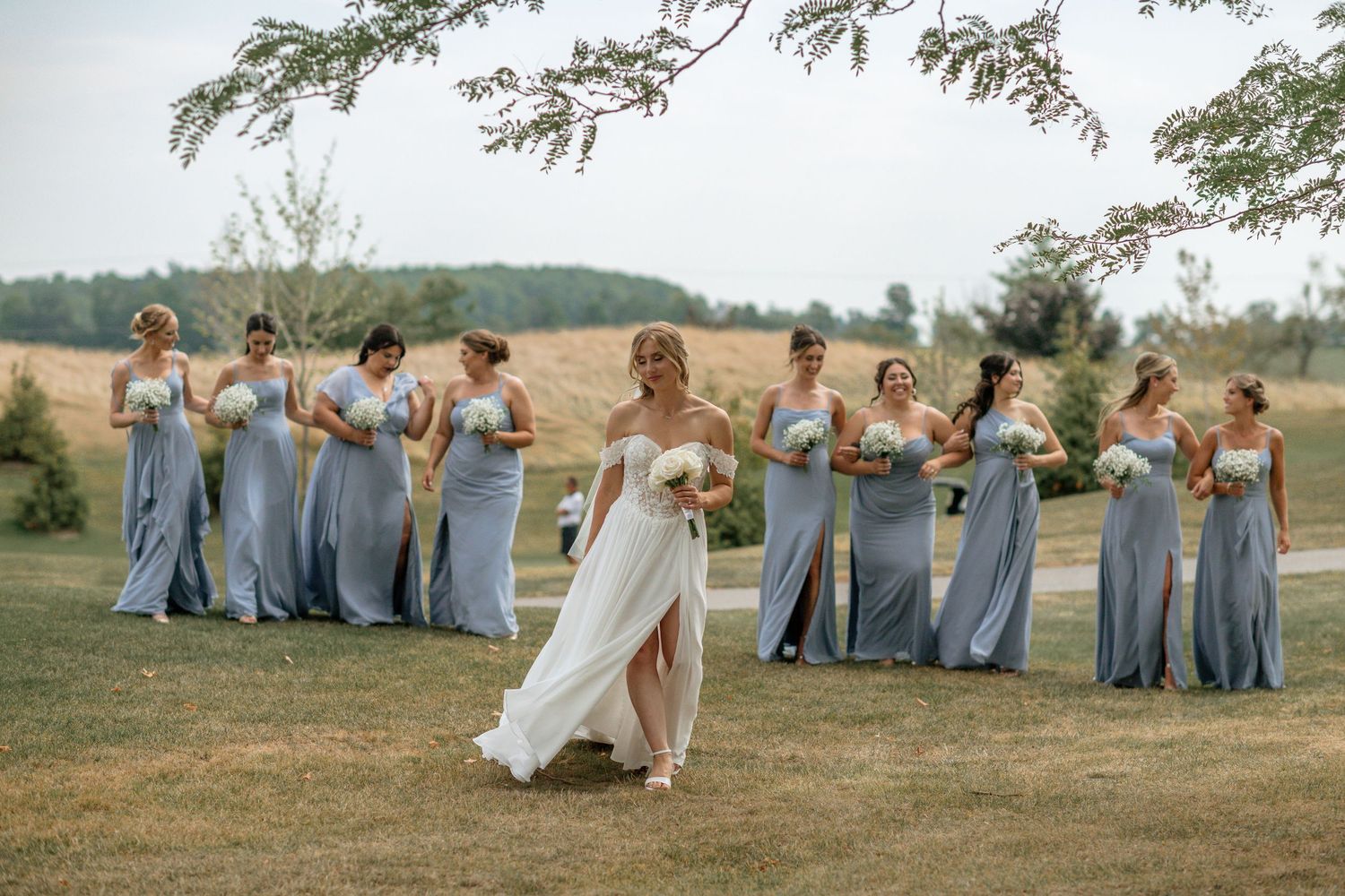 Wedding party in light blue dresses poses playfully on a grassy field during sunset.
