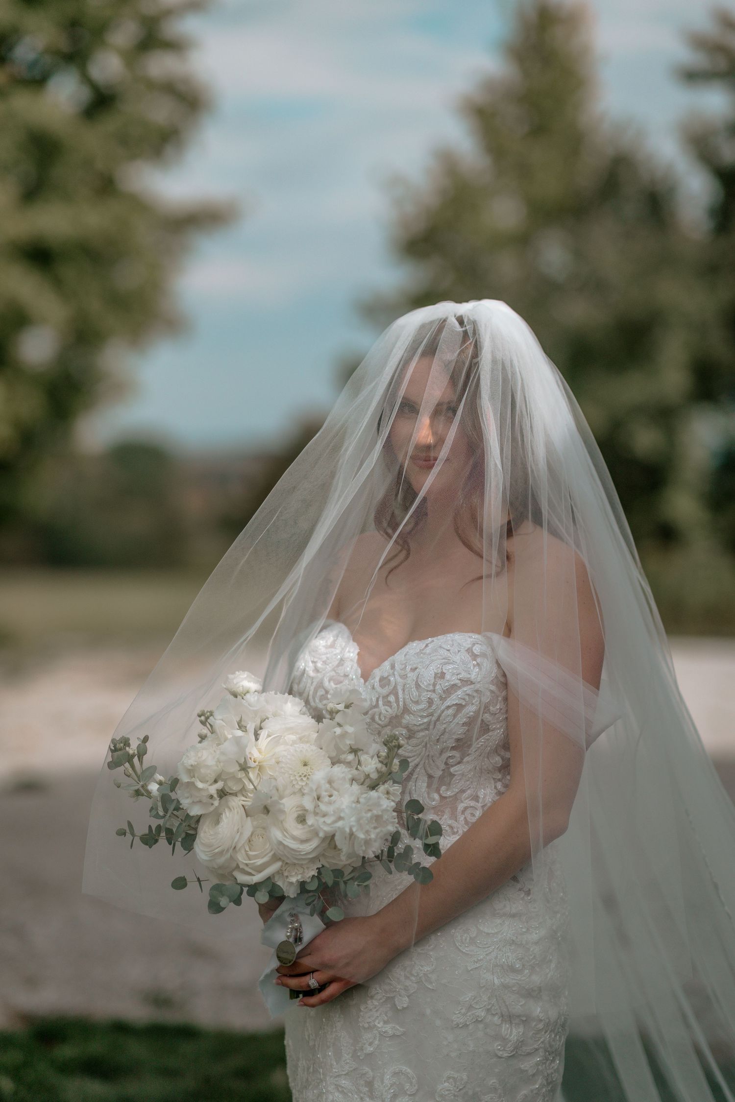 Bride in lace dress with flowing veil holds white and green bouquet outdoors.