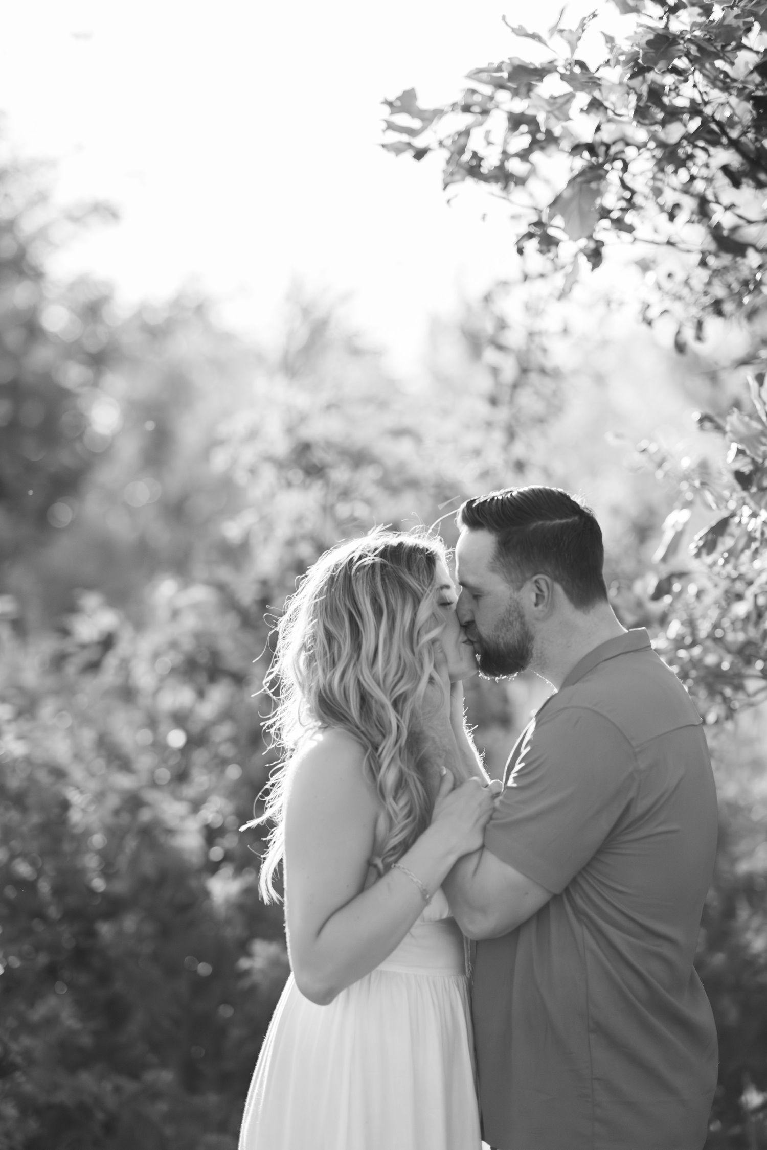 Romantic couple sharing a tender kiss in a sunlit garden with blooming trees in the background.