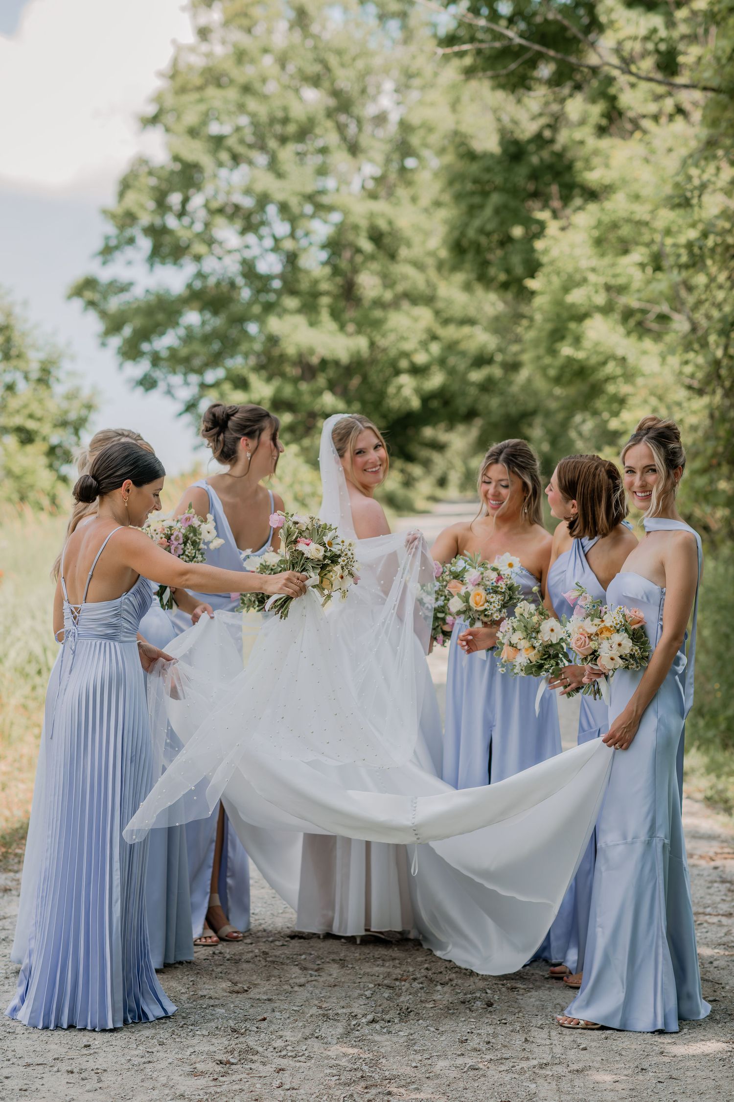 A wedding party in light blue bridesmaid dresses helping arrange the bride's dress outdoors.