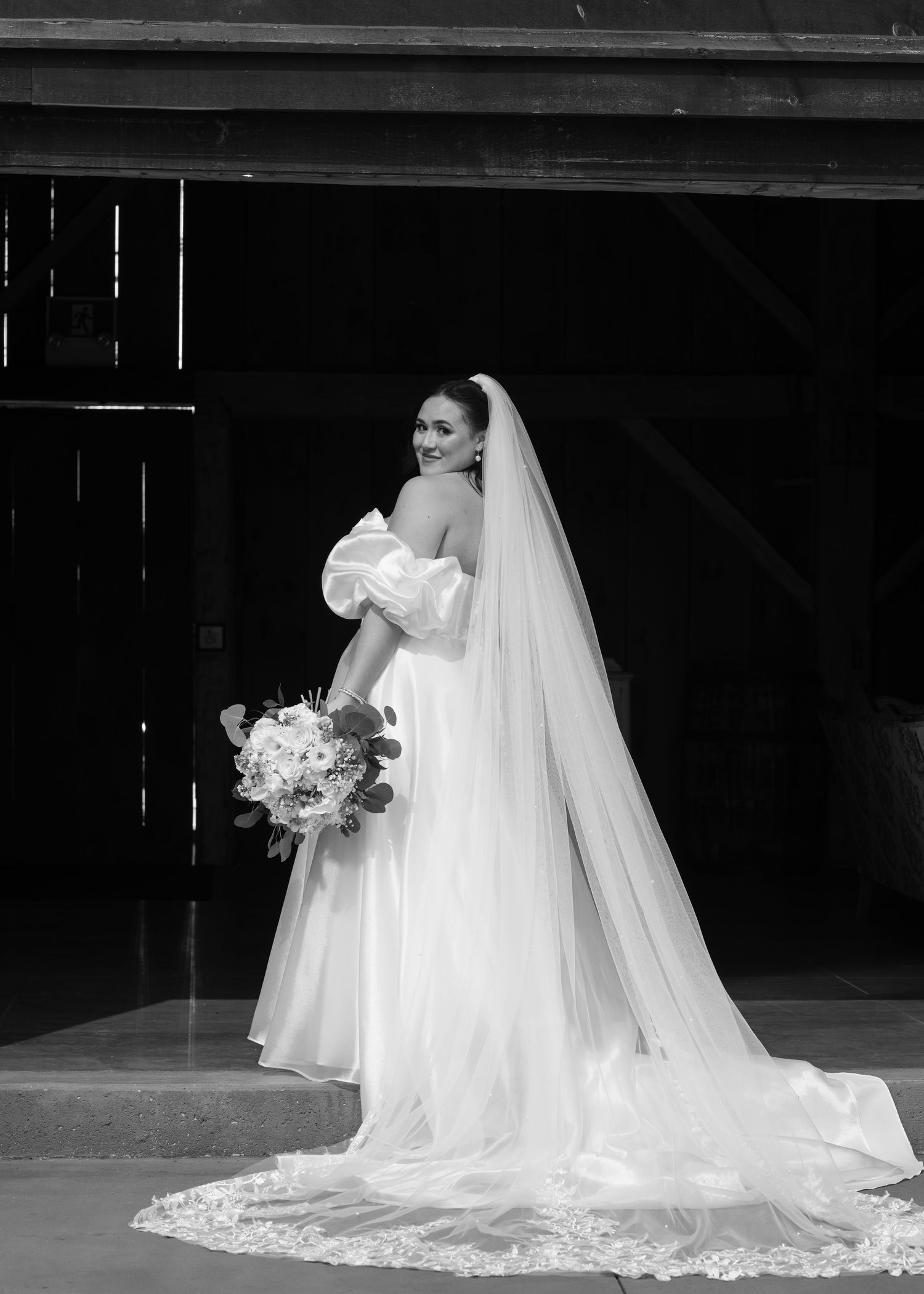 A bride in an elegant white gown with a long lace veil poses in a barn doorway holding a bouquet.