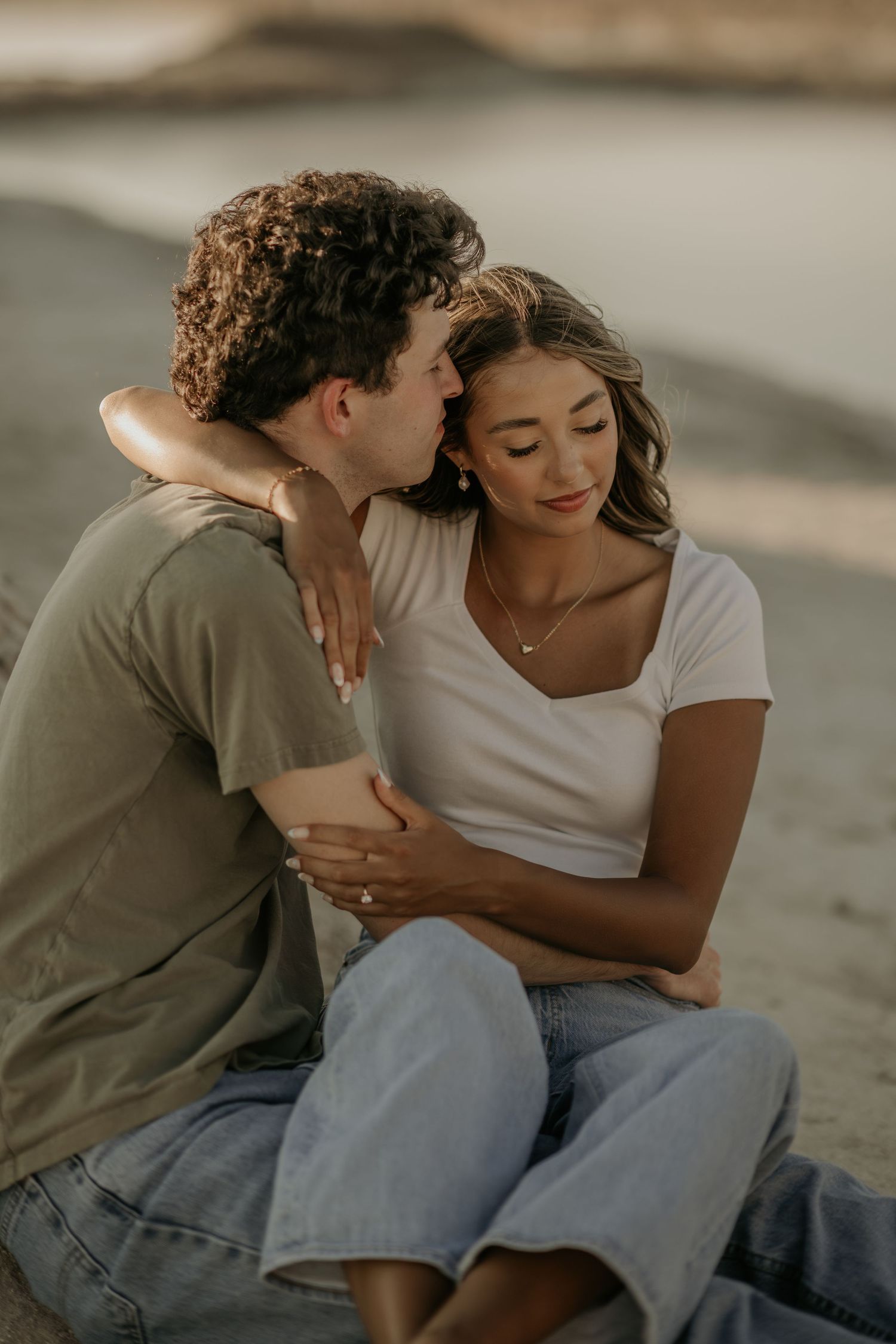 A couple shares intimate moments together while sitting on a beach at sunset in casual attire.