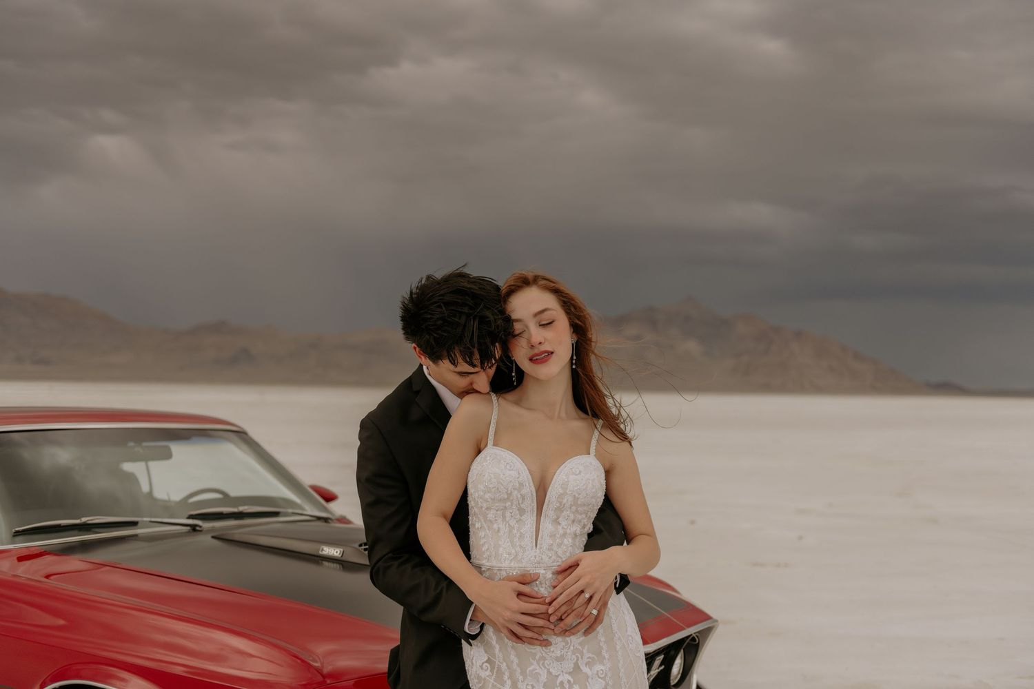 A couple in formal attire embraces on a scenic salt flat with dramatic cloudy skies and mountains in the background.