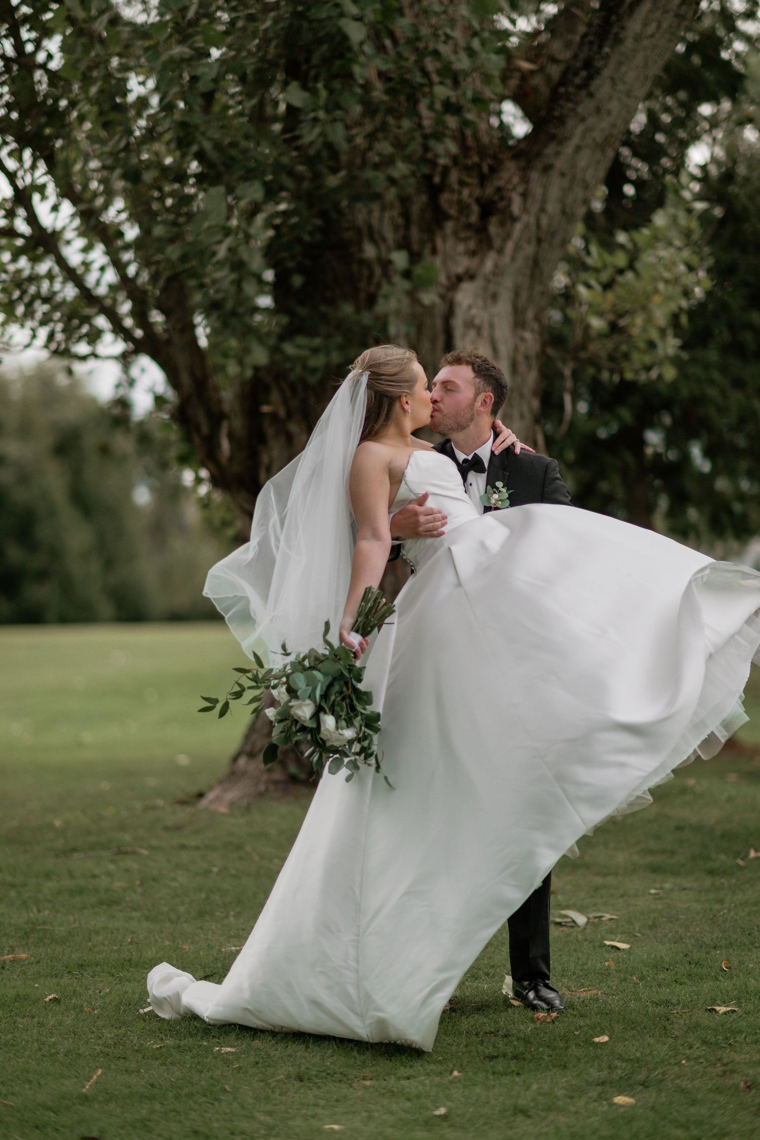 A romantic wedding couple shares a tender moment in a grassy field beneath a large tree.