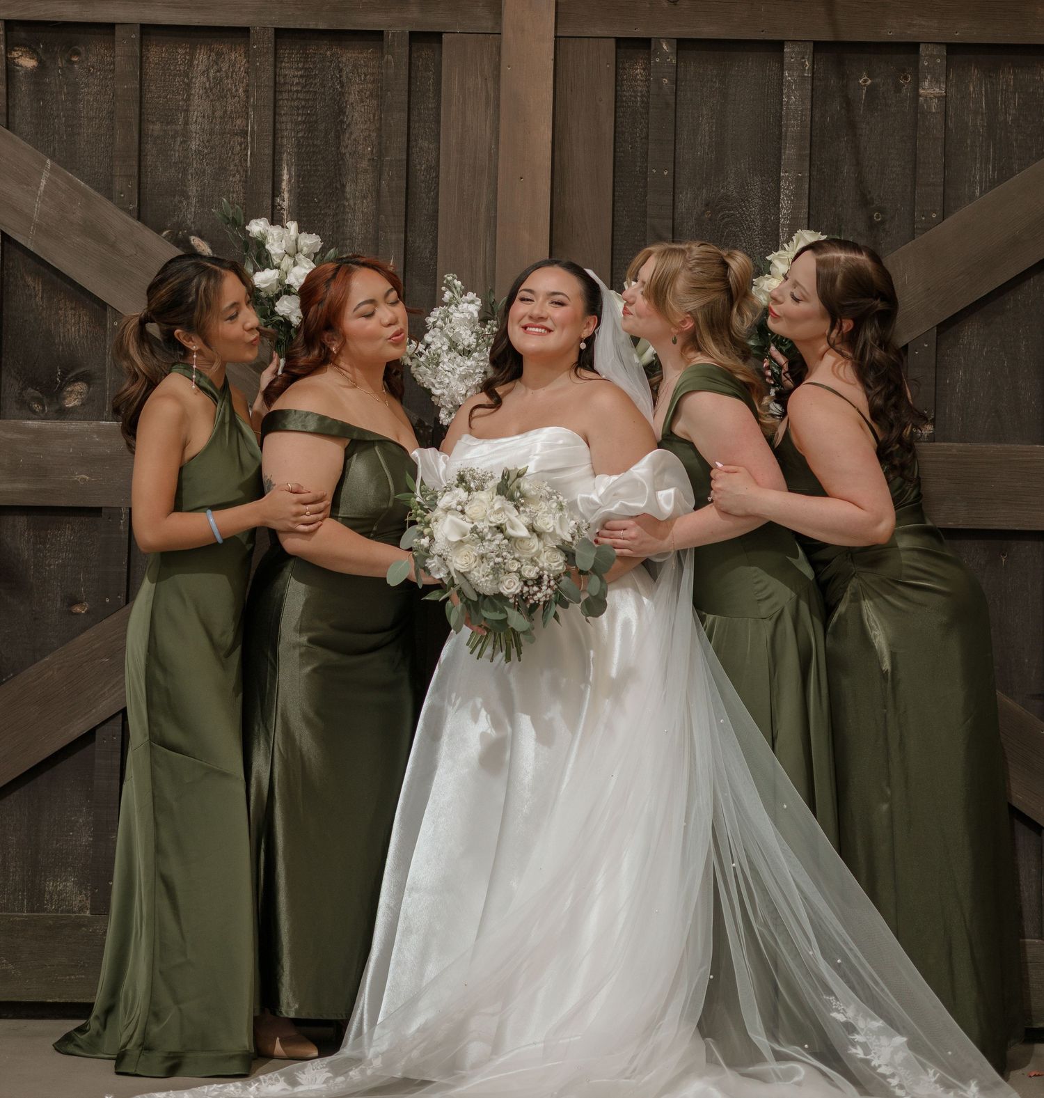 Bridesmaids in olive green dresses share an emotional moment with bride in white gown against rustic wooden backdrop.