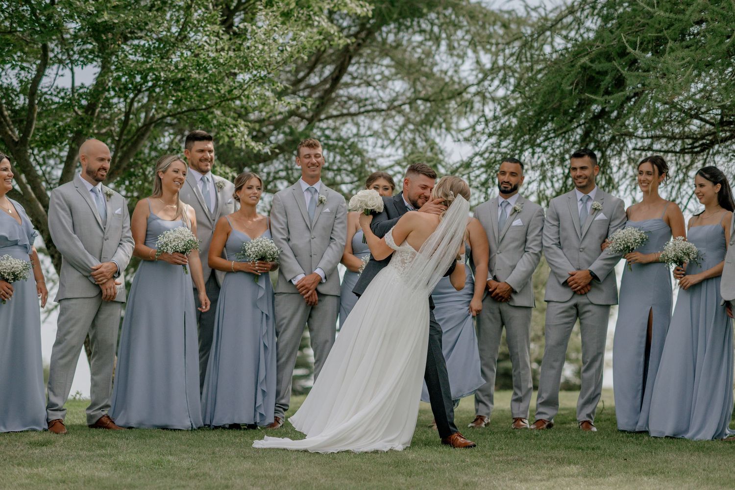 Wedding party in blue dresses and gray suits poses together under trees.