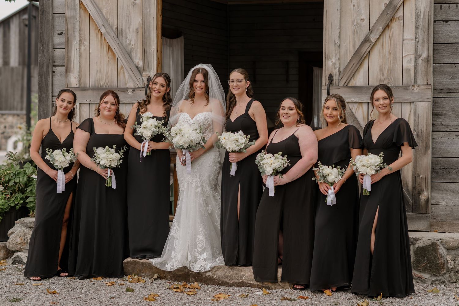 Wedding party in black dresses and white gown pose together against rustic barn backdrop.