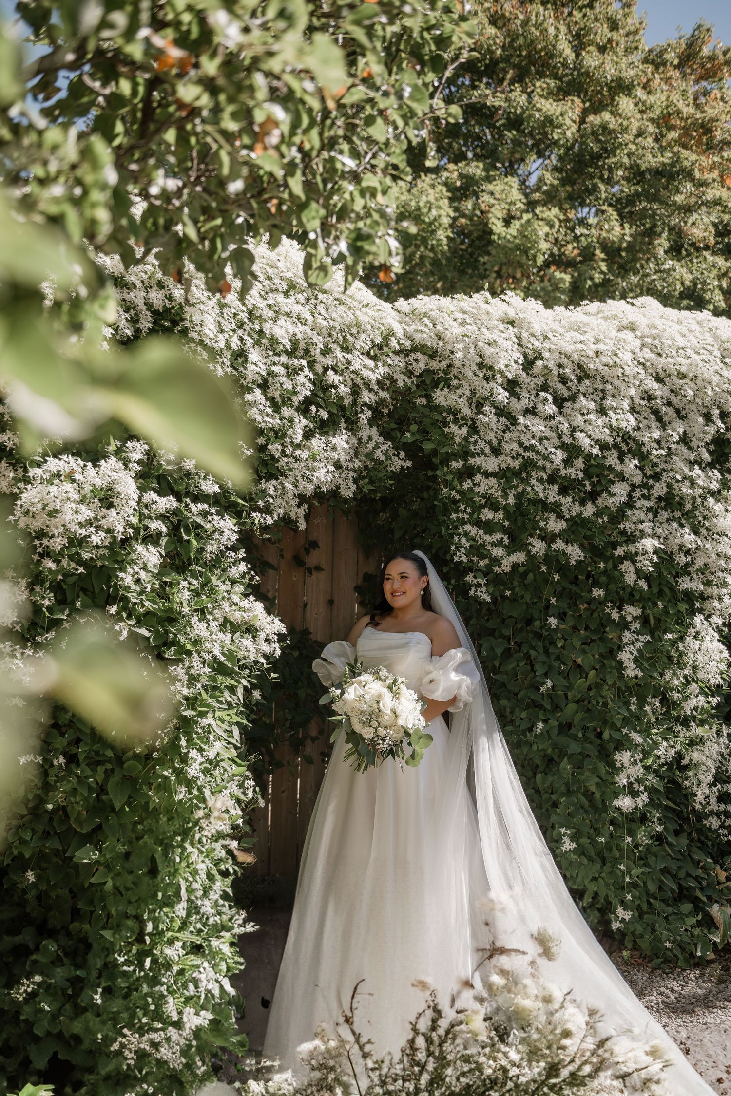 A white wedding dress stands out against a stunning backdrop of climbing white flowers and greenery in a garden setting.