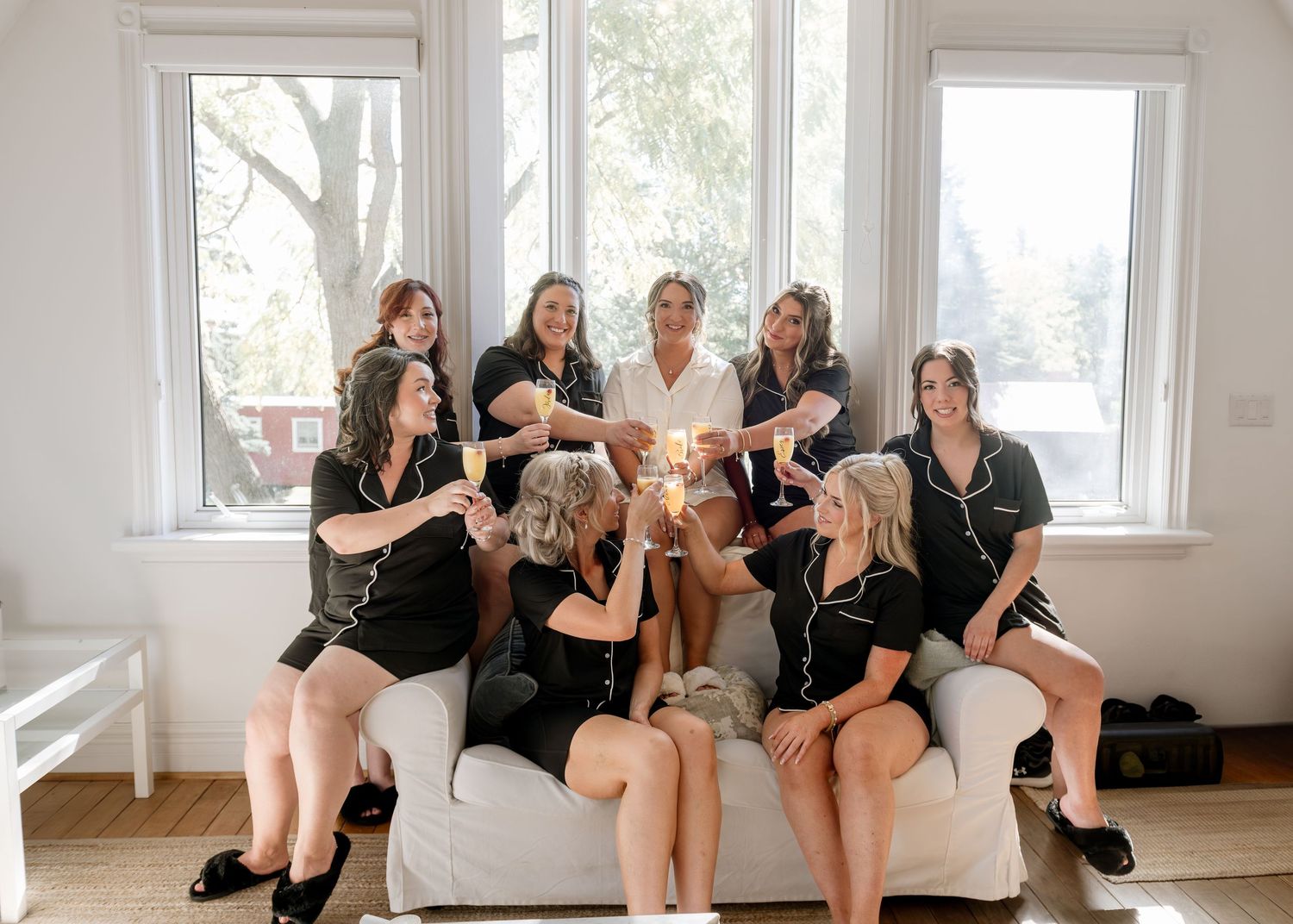 A group of friends in black outfits pose together on a white couch in a bright, sunlit room.