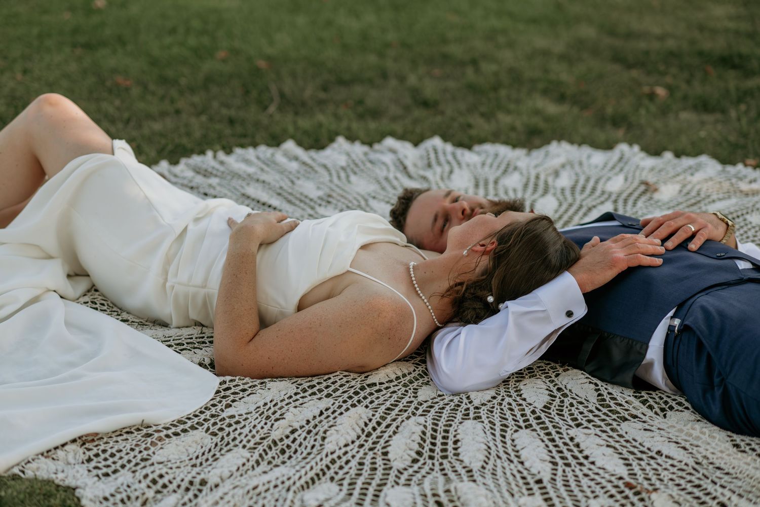 A couple in wedding attire lay together on a white lace blanket in the grass during an outdoor photoshoot.