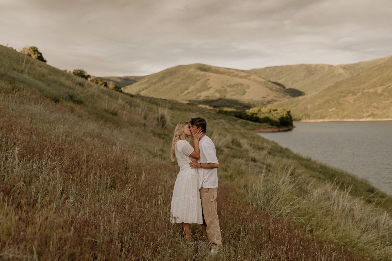 A couple embraces on a hillside overlooking a serene lake and rolling hills during golden hour.