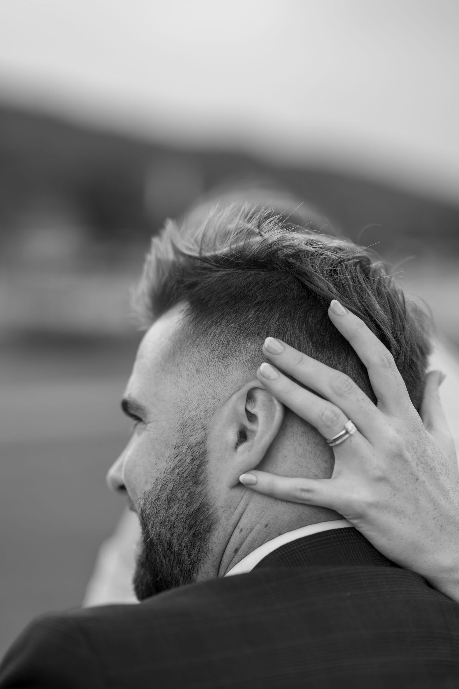Black and white photo showing a hand with a wedding ring touching the back of a person's head with styled hair.