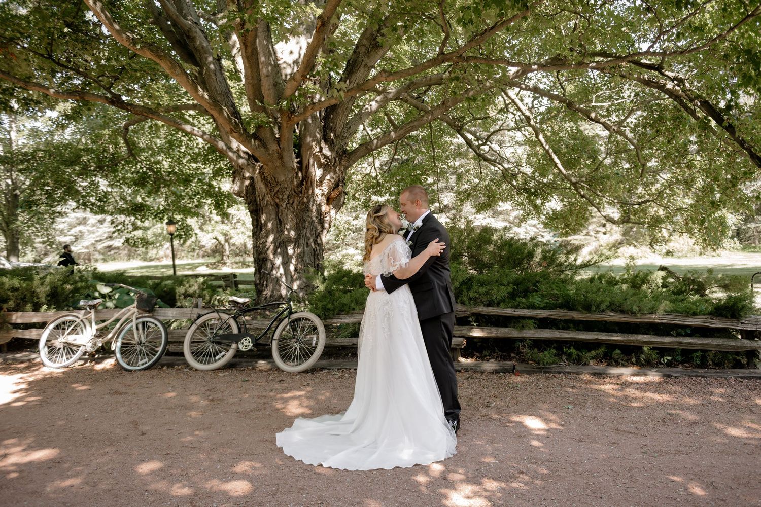 Wedding couple walking together under lush green trees with bicycles parked in the background.