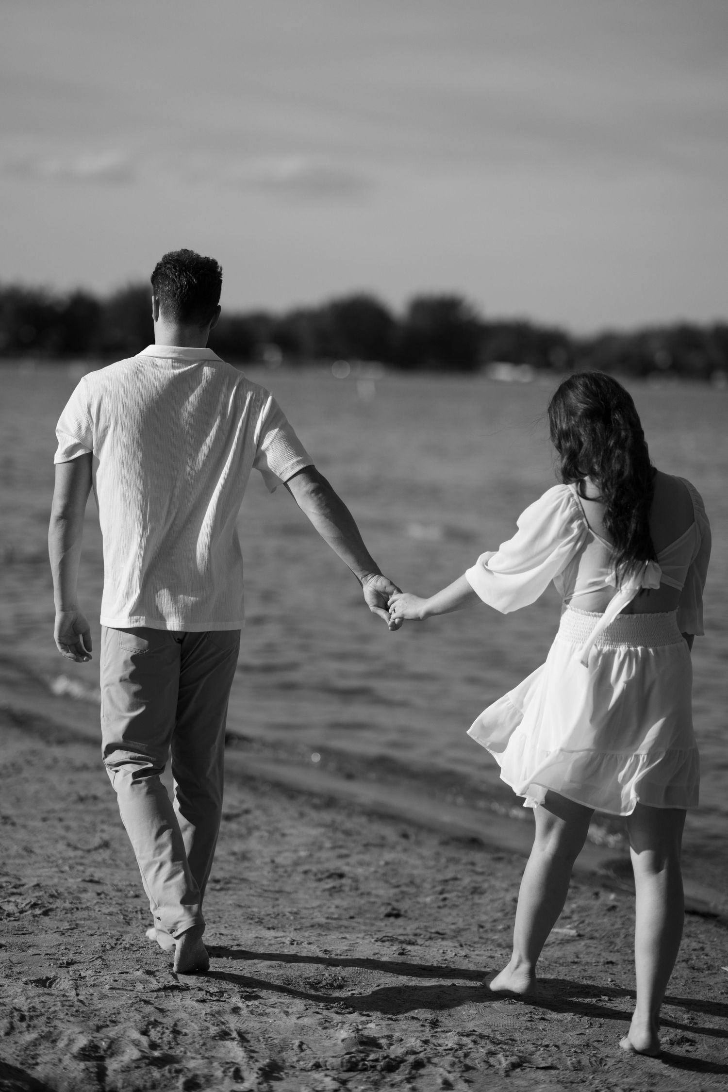 A couple walks hand in hand along a beach shore in black and white.