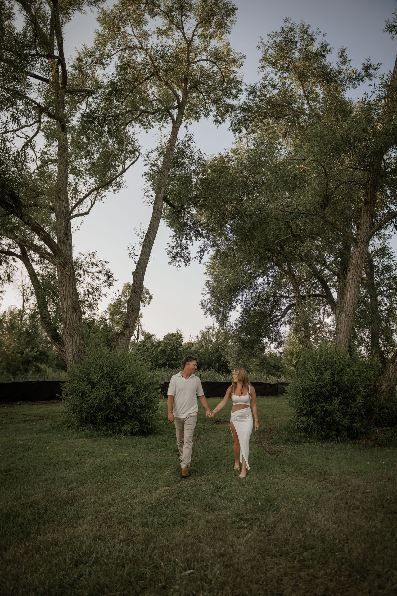 Couple strolls through scenic park with tall trees at dusk.