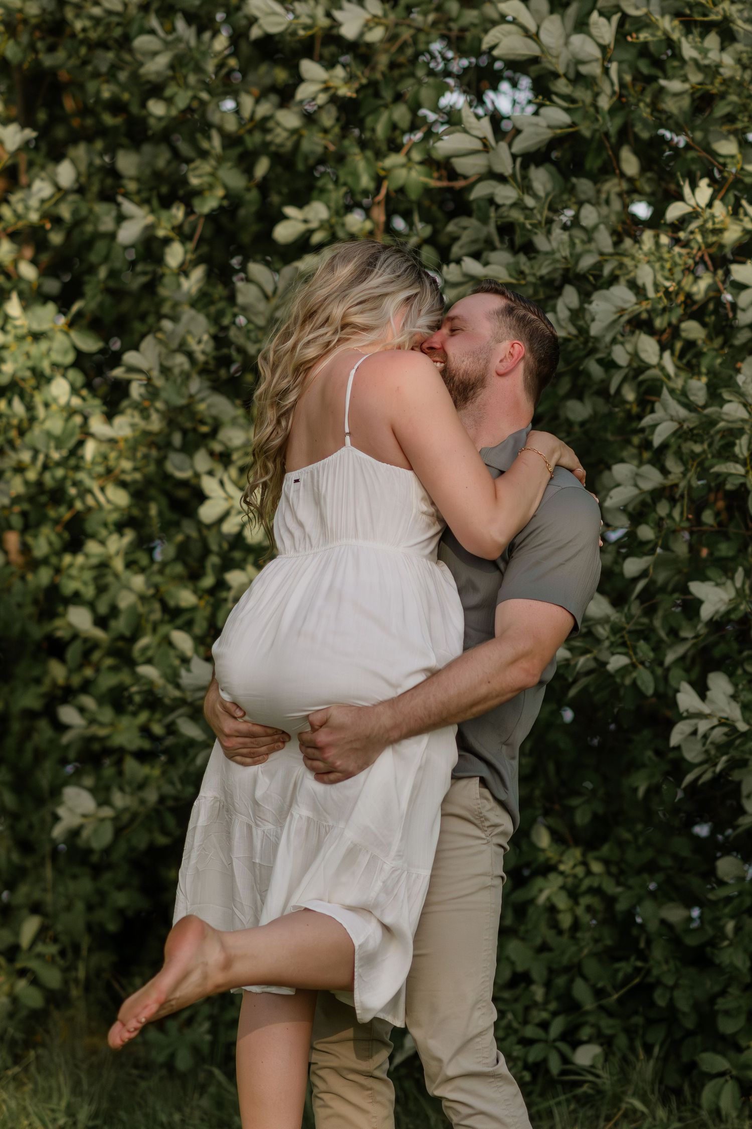Couple in white and khaki spins gracefully on grass near blooming trees.
