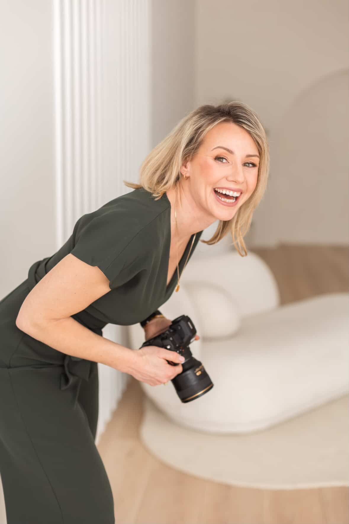 Professional photographer in olive green dress smiles while holding DSLR camera in bright interior setting.