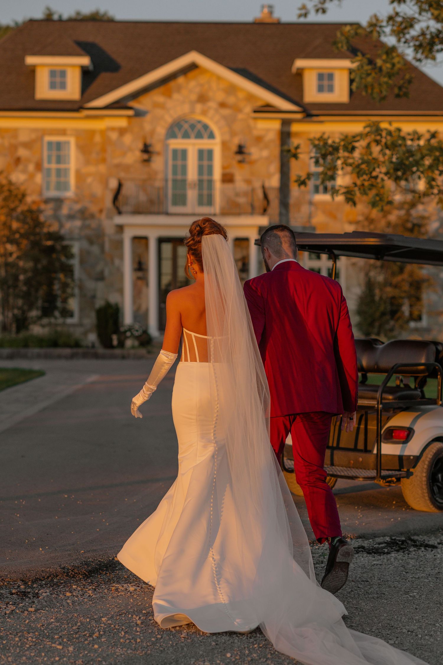 Wedding dress and red suit silhouetted against a stone building at golden hour.