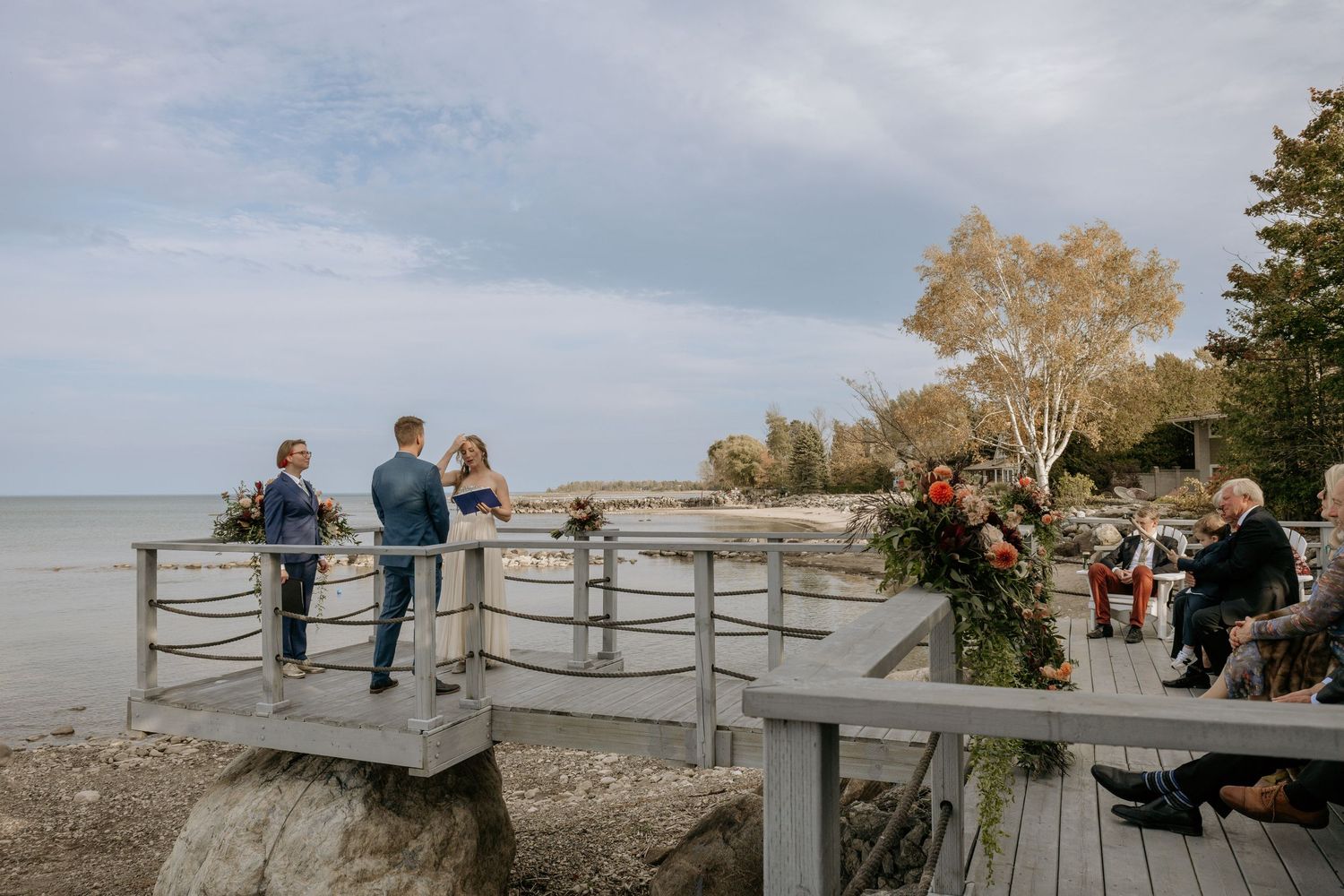People gather on a wooden boardwalk overlooking a serene waterfront on a sunny autumn day.