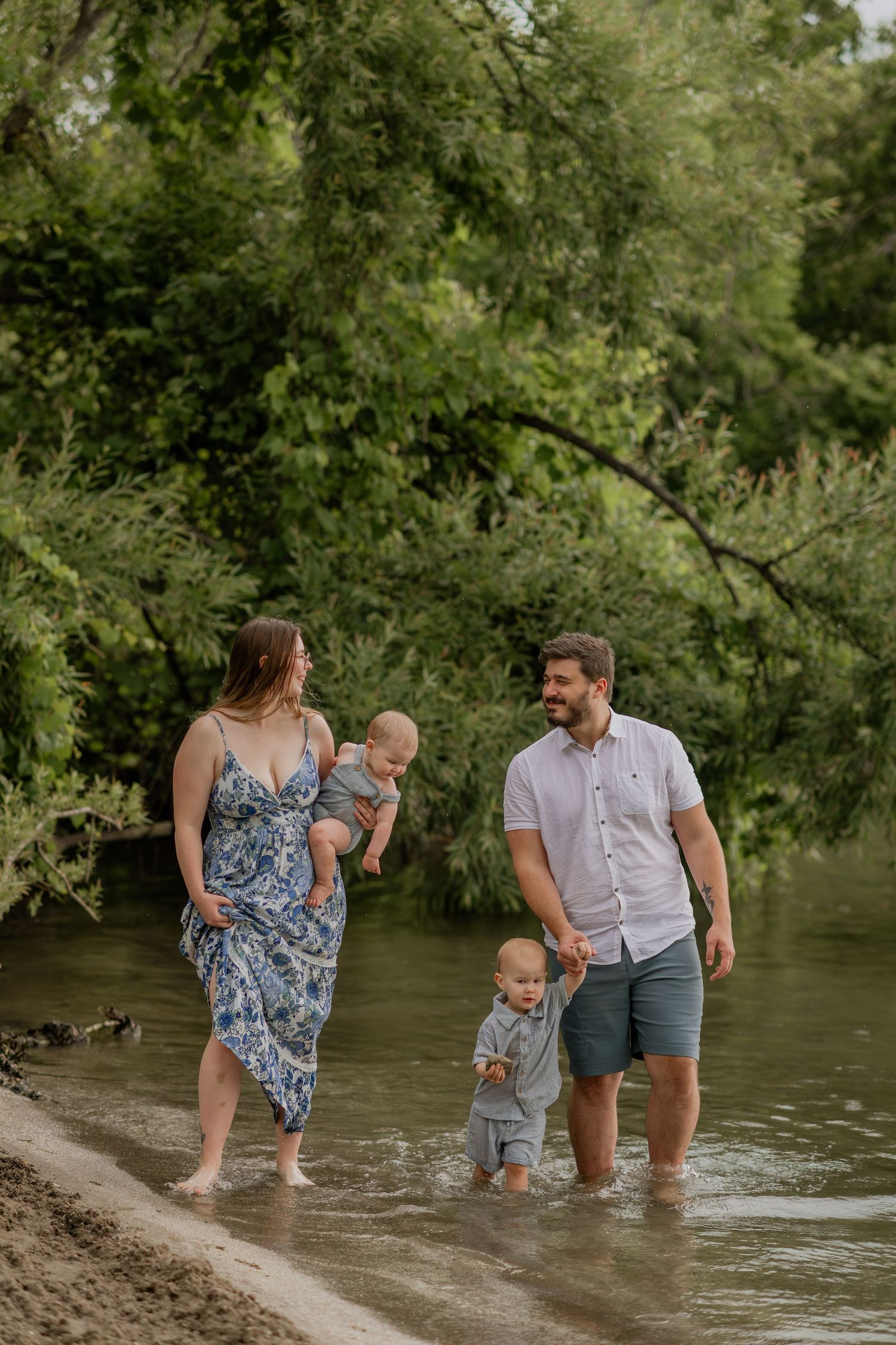 Family wading together in shallow river water surrounded by green trees.