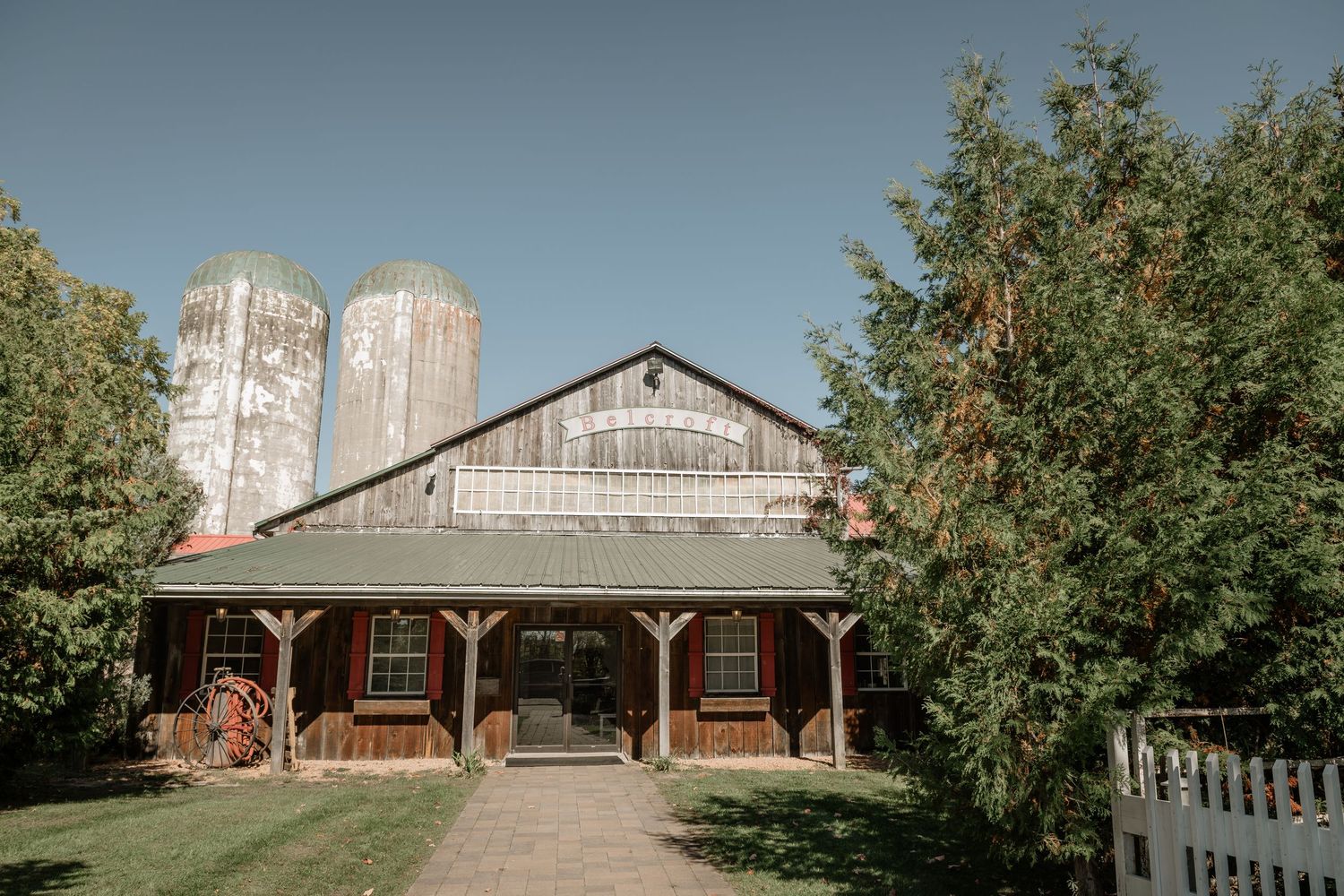 A rustic barn with metal silos stands against a backdrop of green trees on a sunny day.