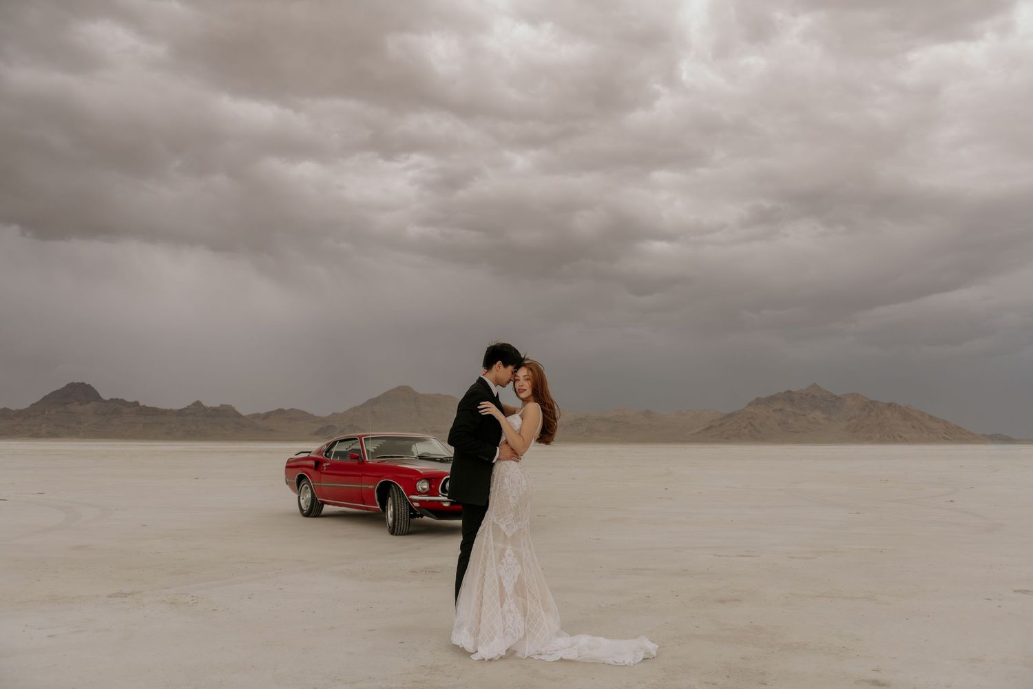 A couple in wedding attire poses dramatically against the stark backdrop of Bonneville Salt Flats under moody storm clouds.