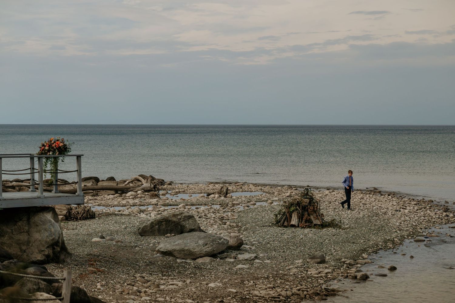 A serene panoramic view of a rocky beach shoreline with calm waters and cloudy skies stretching to the horizon.