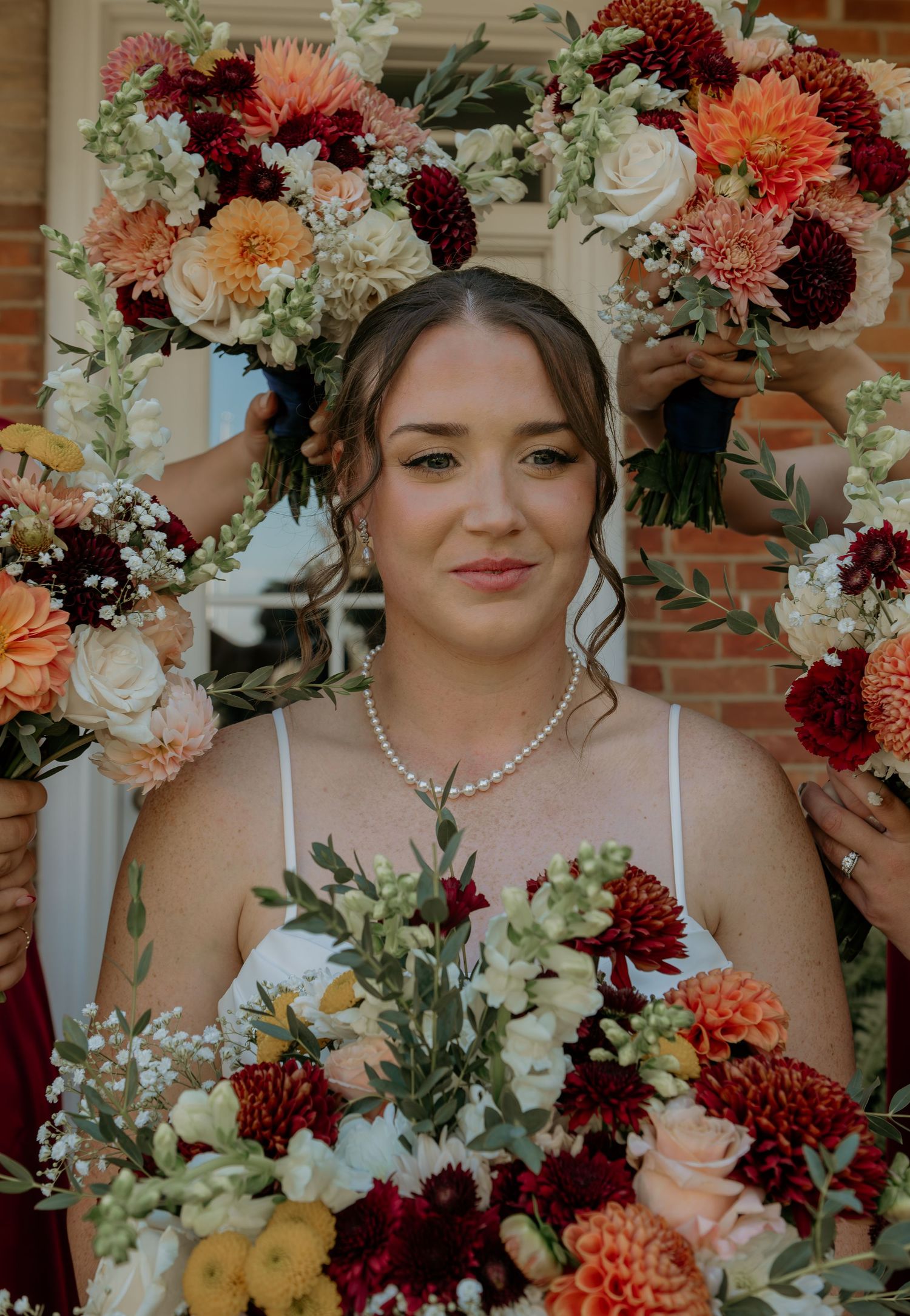 Beautiful autumn wedding bouquets with burgundy dahlias, peach roses, and white hydrangeas frame a pearl-adorned bride.