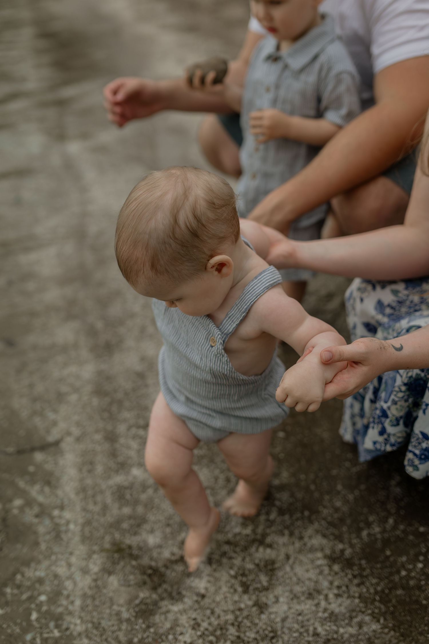 Baby learning to walk on beach with supporting hands guiding their steps.