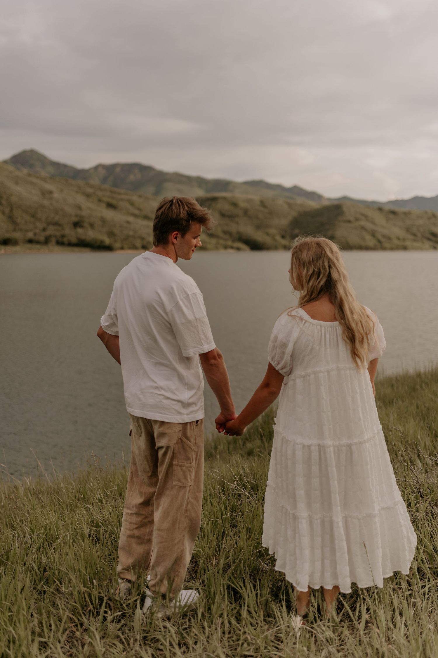 Couple in white and beige outfits holding hands while walking through tall grass near a scenic mountain lake.