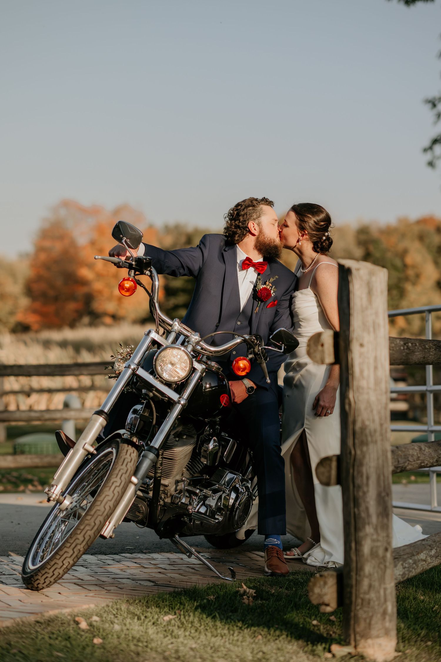 A romantic motorcycle engagement photo shoot at sunset with a couple posing near wooden fence posts in autumn.
